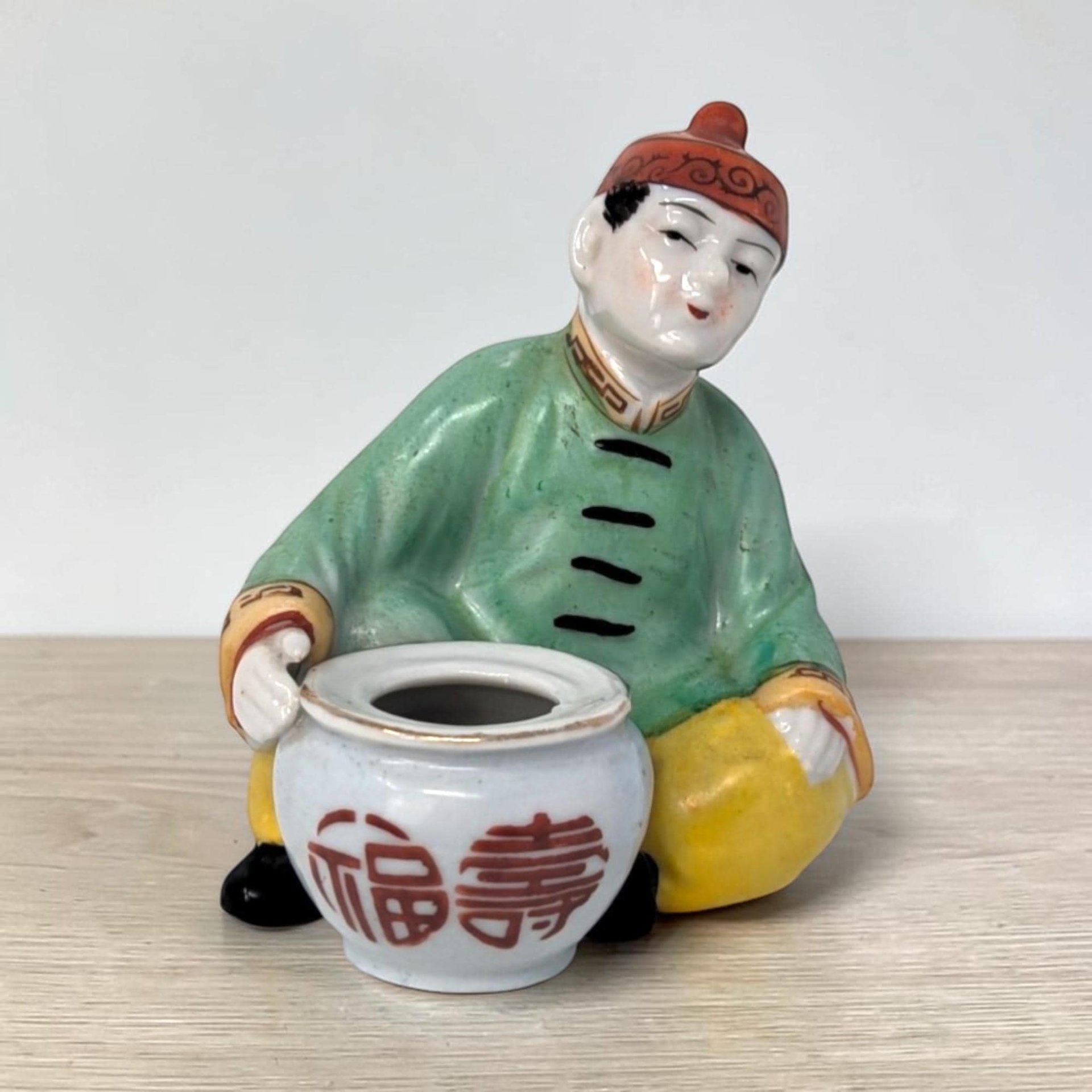 A porcelain figurine of a seated person in a green robe and red cap leaning over a small white pot. The pot features red calligraphic characters, and the figure is set against a plain background on a light wood surface.