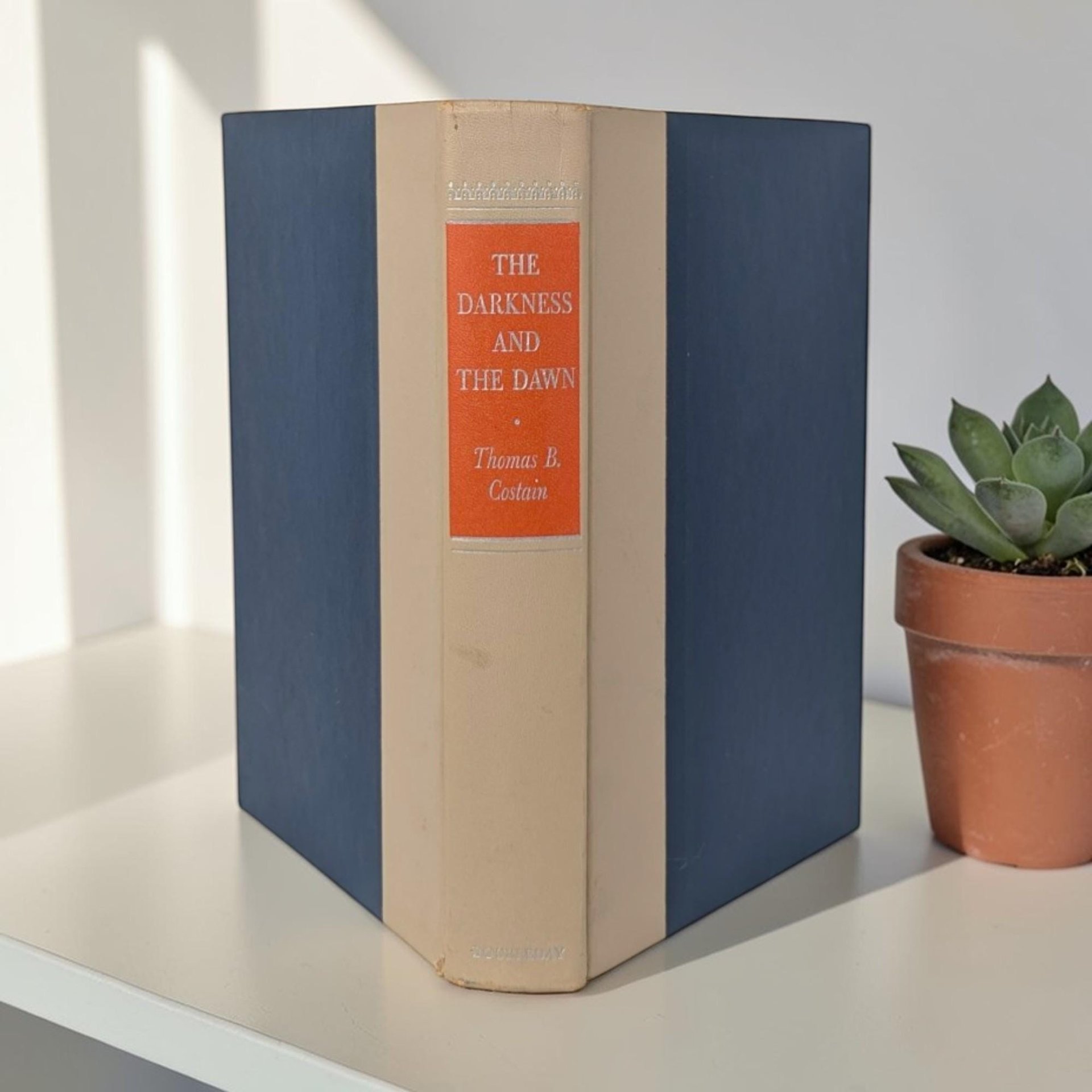 A blue hardcover book with a textured cloth binding. The spine features a cream-stamped title and author name set against an orange black rectangular block.