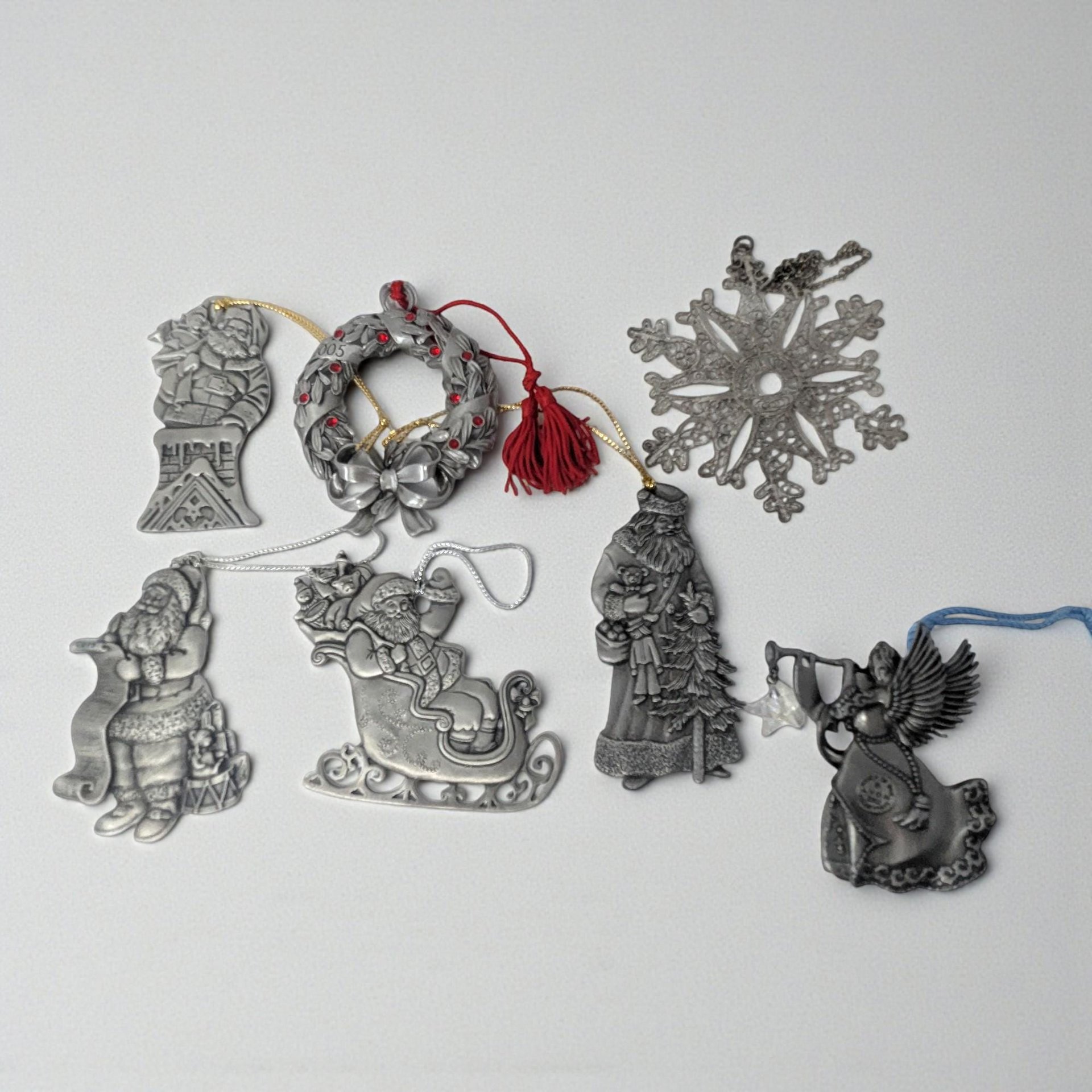 A collection of seven vintage metal ornaments. The set includes detailed pewter figures of Santa in various poses, an angel with a horn, a red-accented wreath with a tassel, and a delicate silver-plated snowflake with an openwork design.