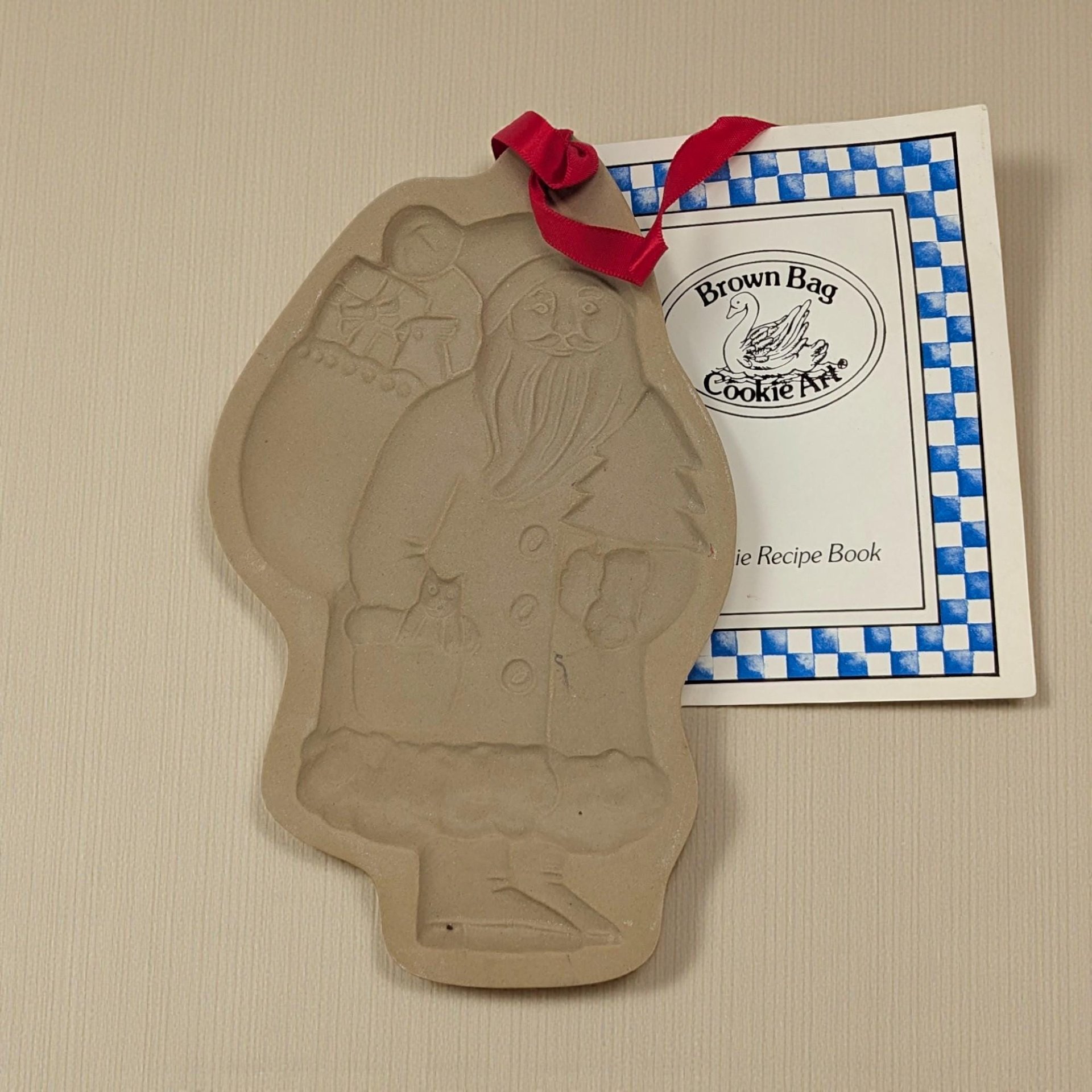 A rectangular stoneware cookie mold featuring a deeply embossed design of a traditional, full-length Santa Claus. He is depicted in a long robe, carrying a sack of toys over his shoulder and a small evergreen tree in his hand, rendered in a warm tan clay.