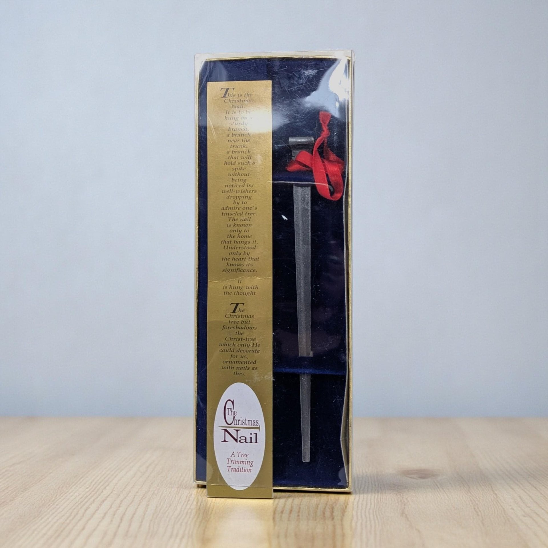 A long, slender metal nail ornament with a textured, dark pewter-toned finish. It features a tapered point and a square head, suspended by a simple red satin ribbon.