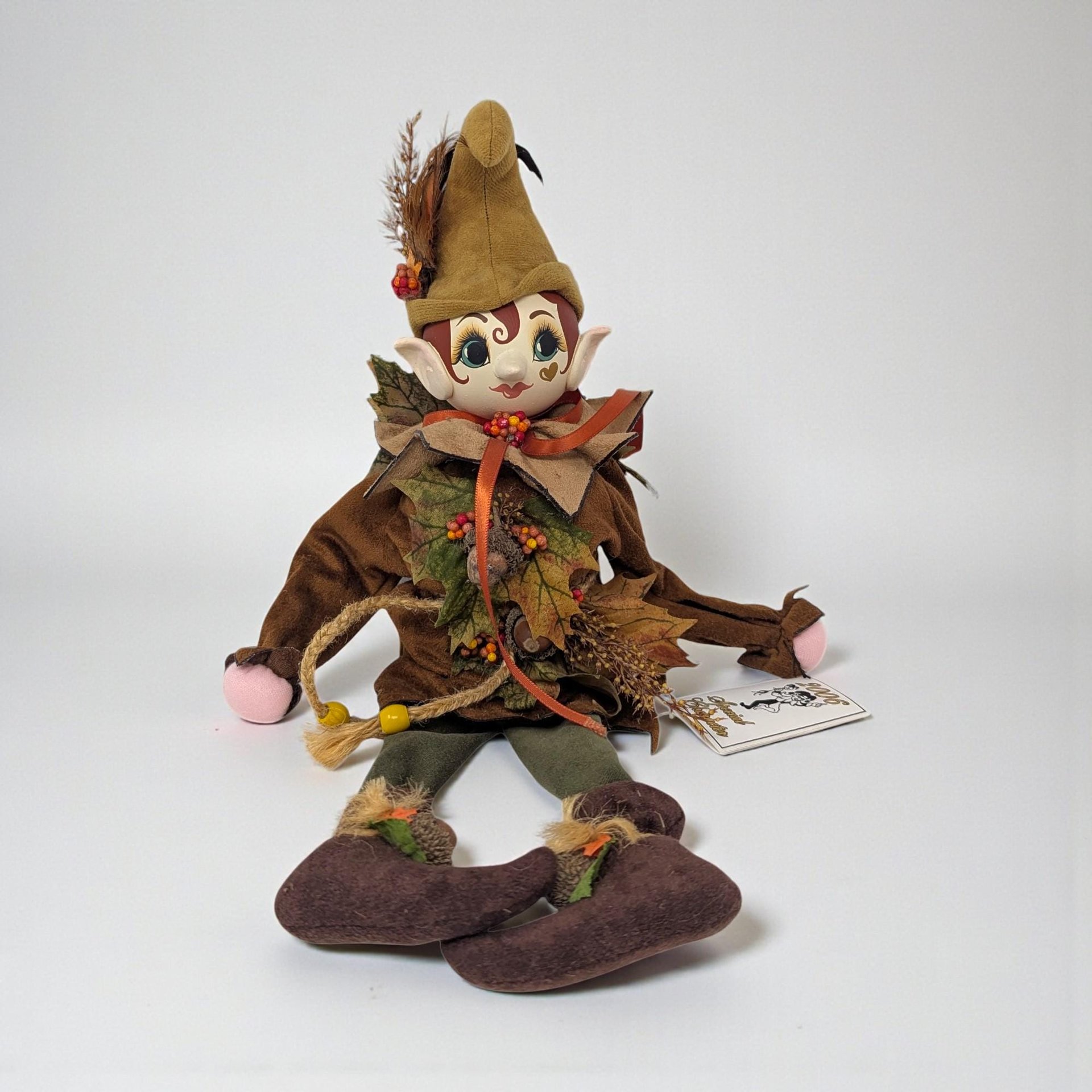 A seated woodland nymph doll with a hand-carved wooden face and soft, fabric hands. She wears a rustic forest outfit in mossy green and brown tones, featuring long, unfilled legs designed to drape over a shelf edge.