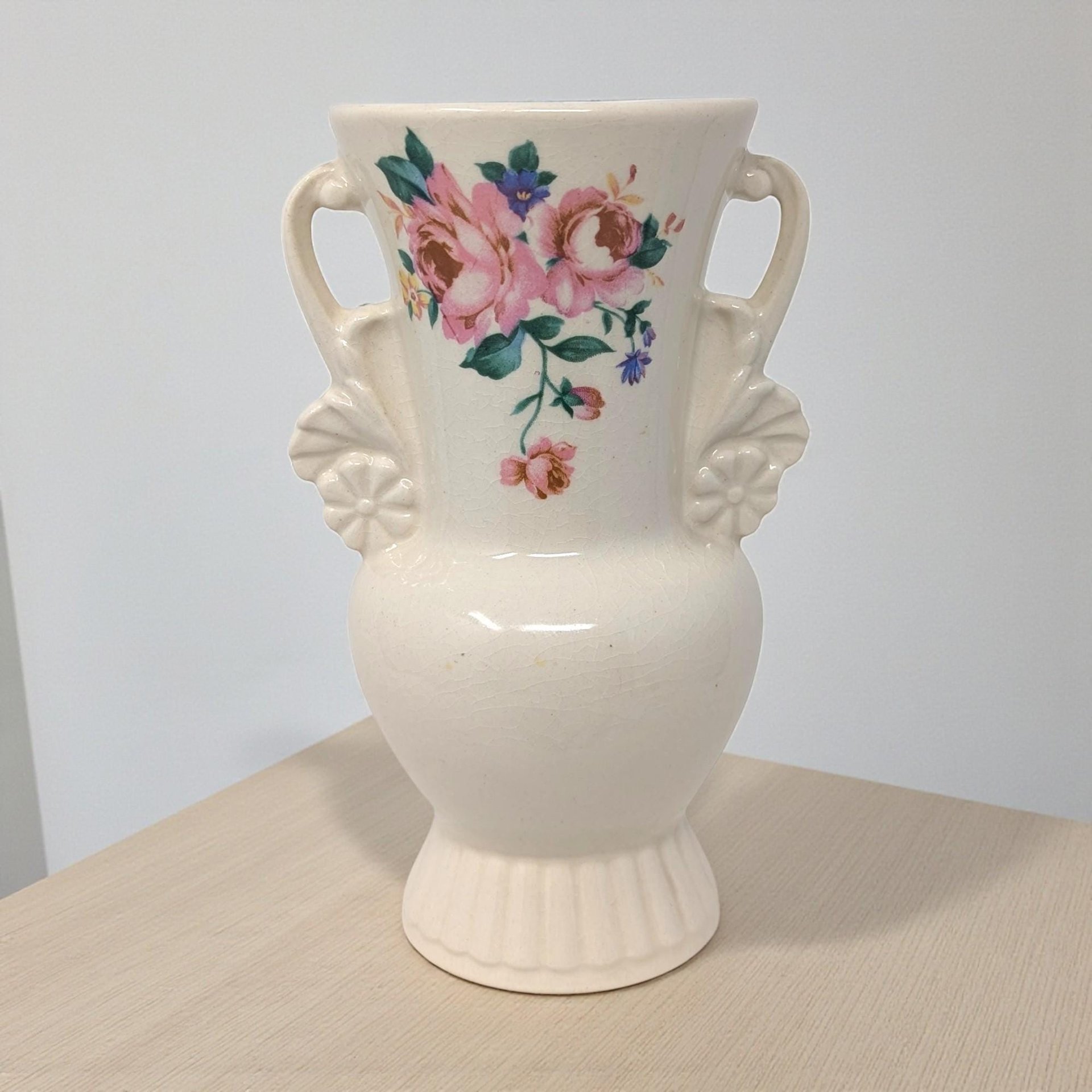 A cream-colored ceramic vase with a flared rim and a round body. It features a raised, sculptural floral motif on the front, finished in a high-gloss glaze with a fine network of surface crazing.