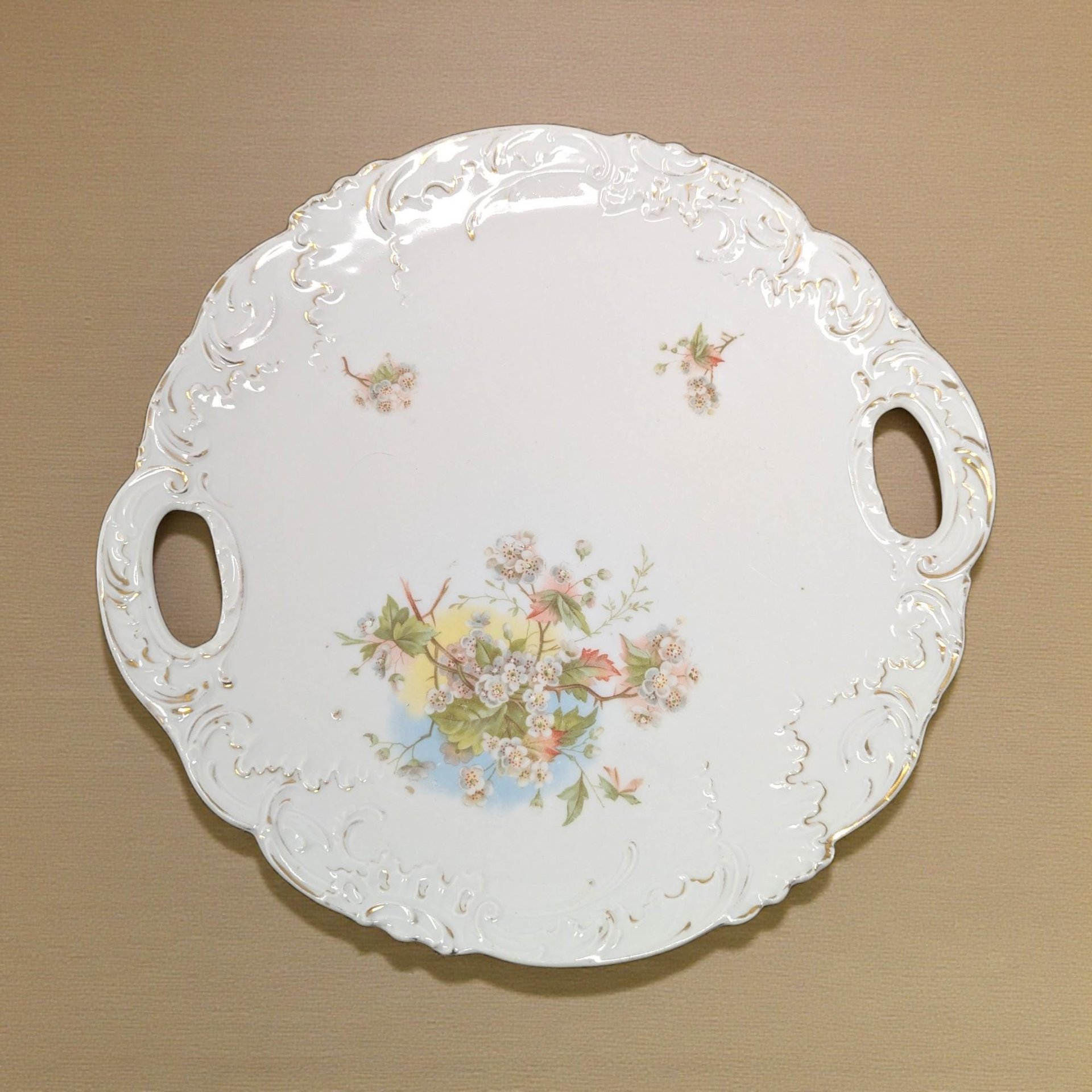 A round white ceramic serving plate with two integrated tab handles. The center features a large multi-colored floral spray, and the scalloped rim shows aged gold gilt accents with visible wear.