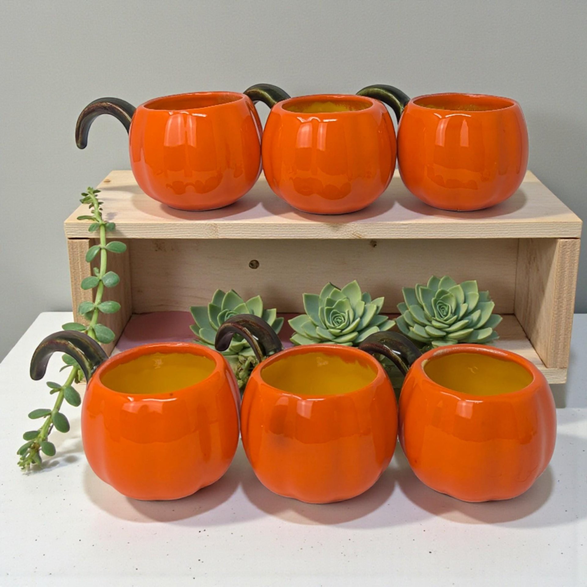 A set of six small ceramic cups shaped like plump orange pumpkins. Each features a green vine-style handle, textured vertical ribbing, and a glossy hand-painted finish.