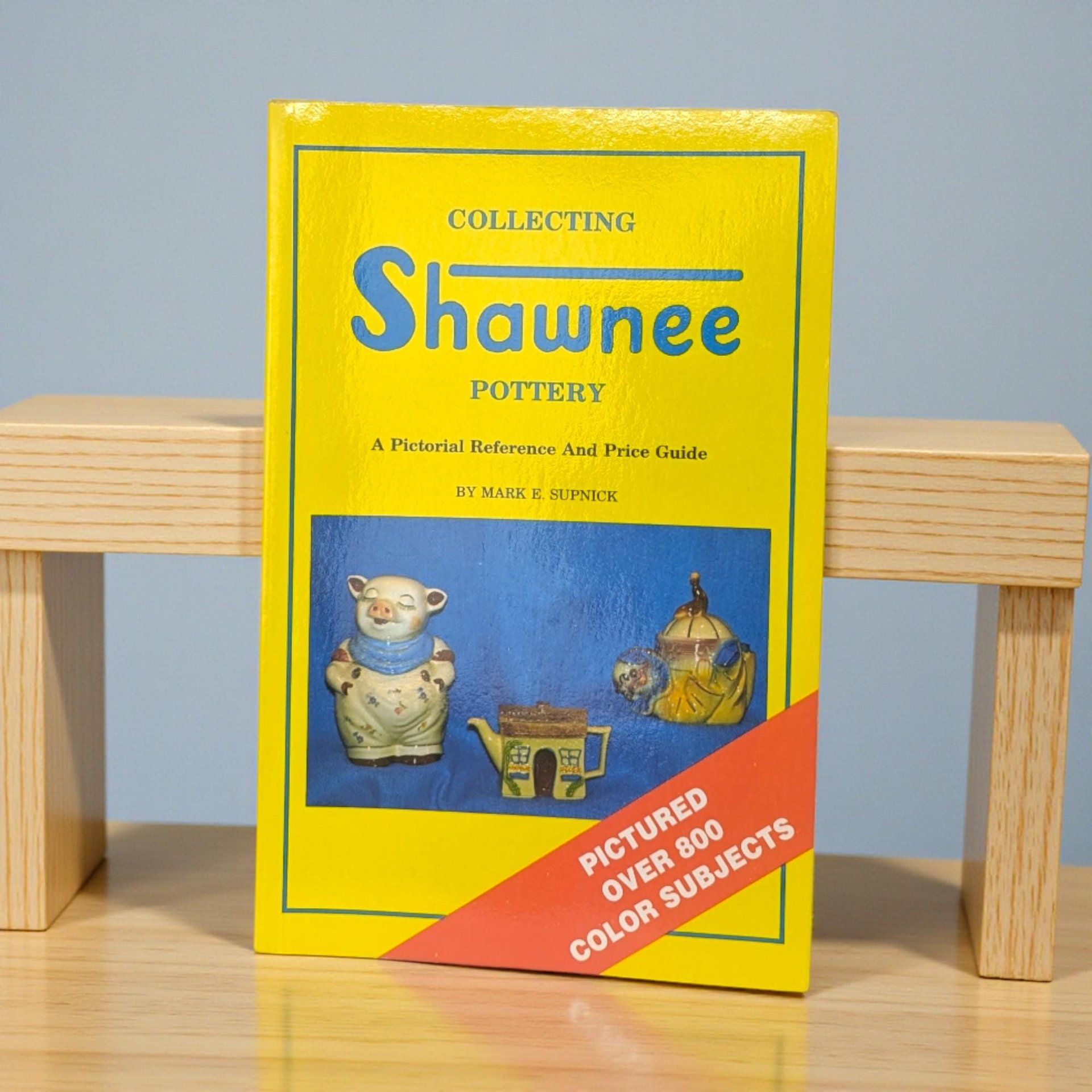 A softcover reference book with a bright yellow cover featuring photographs of various Shawnee pottery pieces, including a teapot and character jars. The text is bold and easy to read, and the binding is clean.