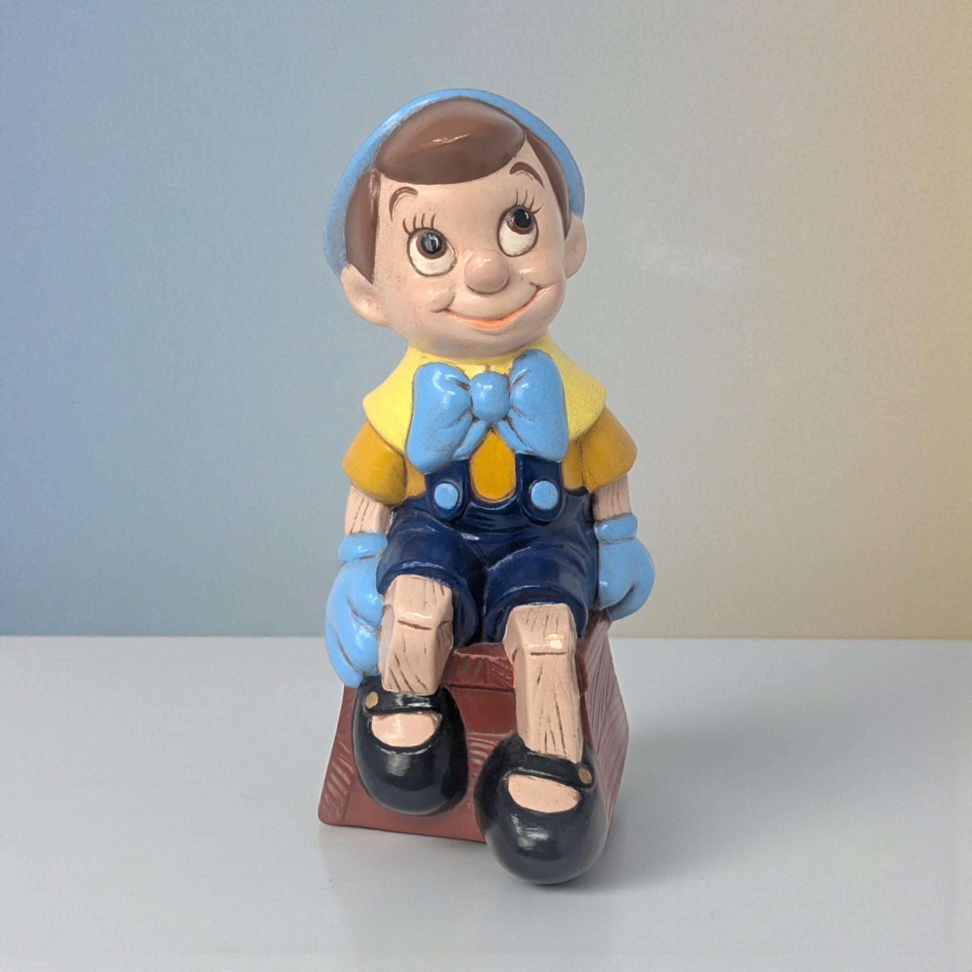 A hand-painted ceramic figure of Pinocchio with a yellow hat, red shorts, and a bright blue bow tie. He stands with an inquisitive expression, his wood-grain limb details subtly rendered in the hand-painted finish.