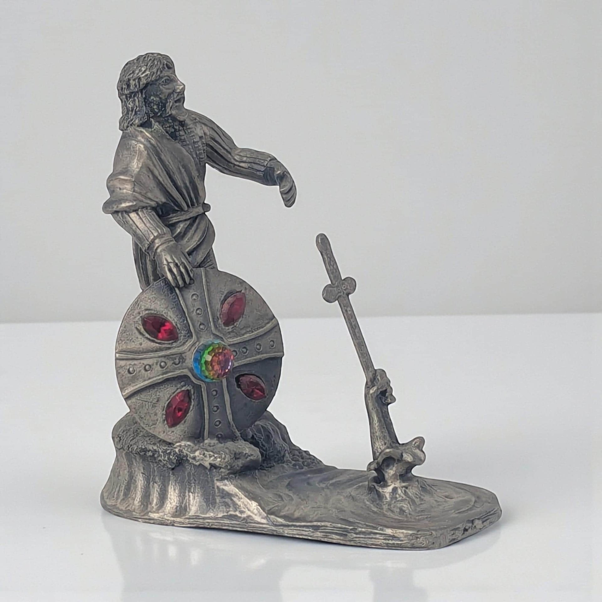 A solid pewter figurine featuring a hand emerging from a swirling pool of water, grasping a detailed sword. The metal has a soft, matte grey finish with intricate textures on the water’s surface and the sword’s hilt.