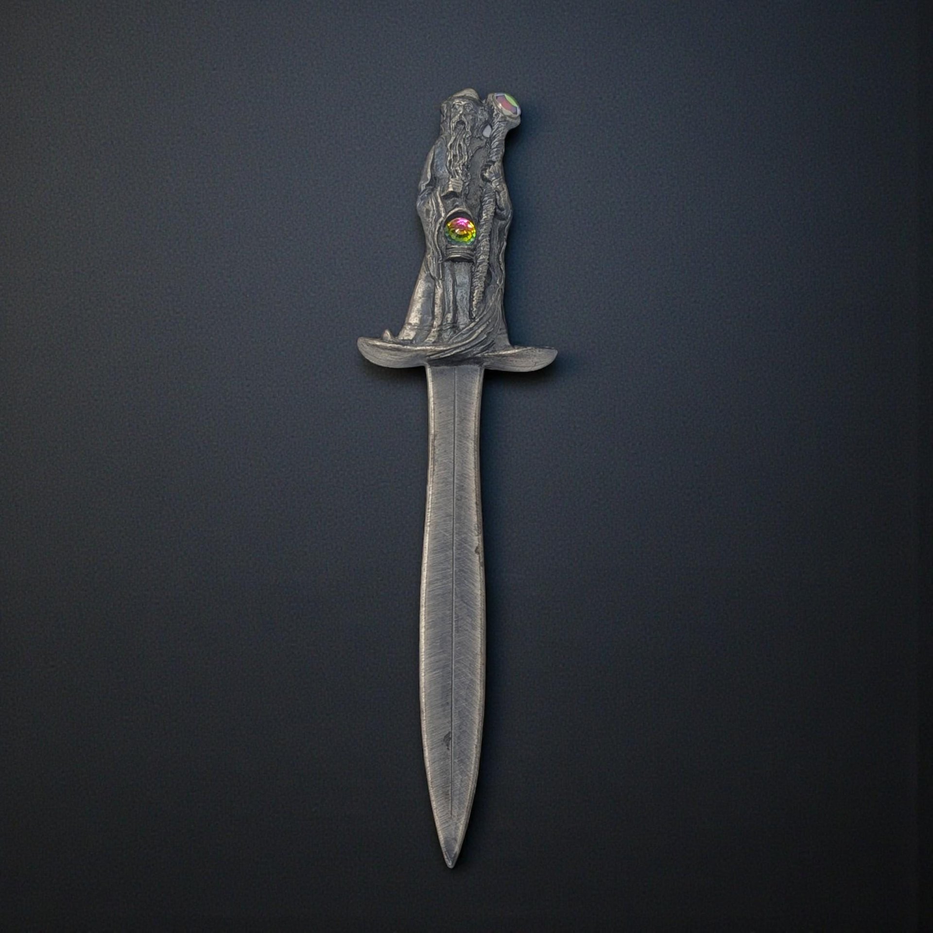 A tall pewter letter opener featuring a multi-towered medieval castle handle topped with a robed wizard figure. The pewter has a dark, antiqued patina that highlights the stonework of the fortress and the flowing robes of the sorcerer.