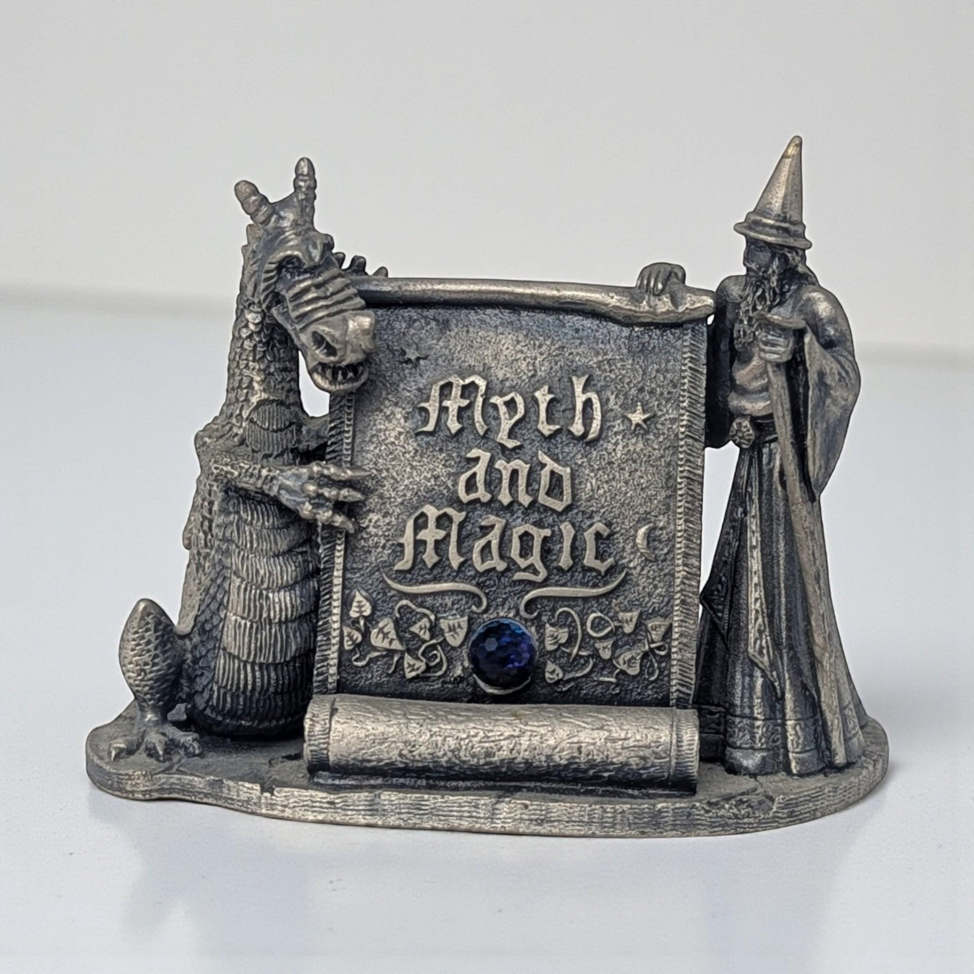 A detailed pewter figurine featuring a robed wizard and a dragon standing on opposite sides of a rustic stone signpost. The base is textured like a rocky path, and the entire piece has a dark, antiqued pewter finish.