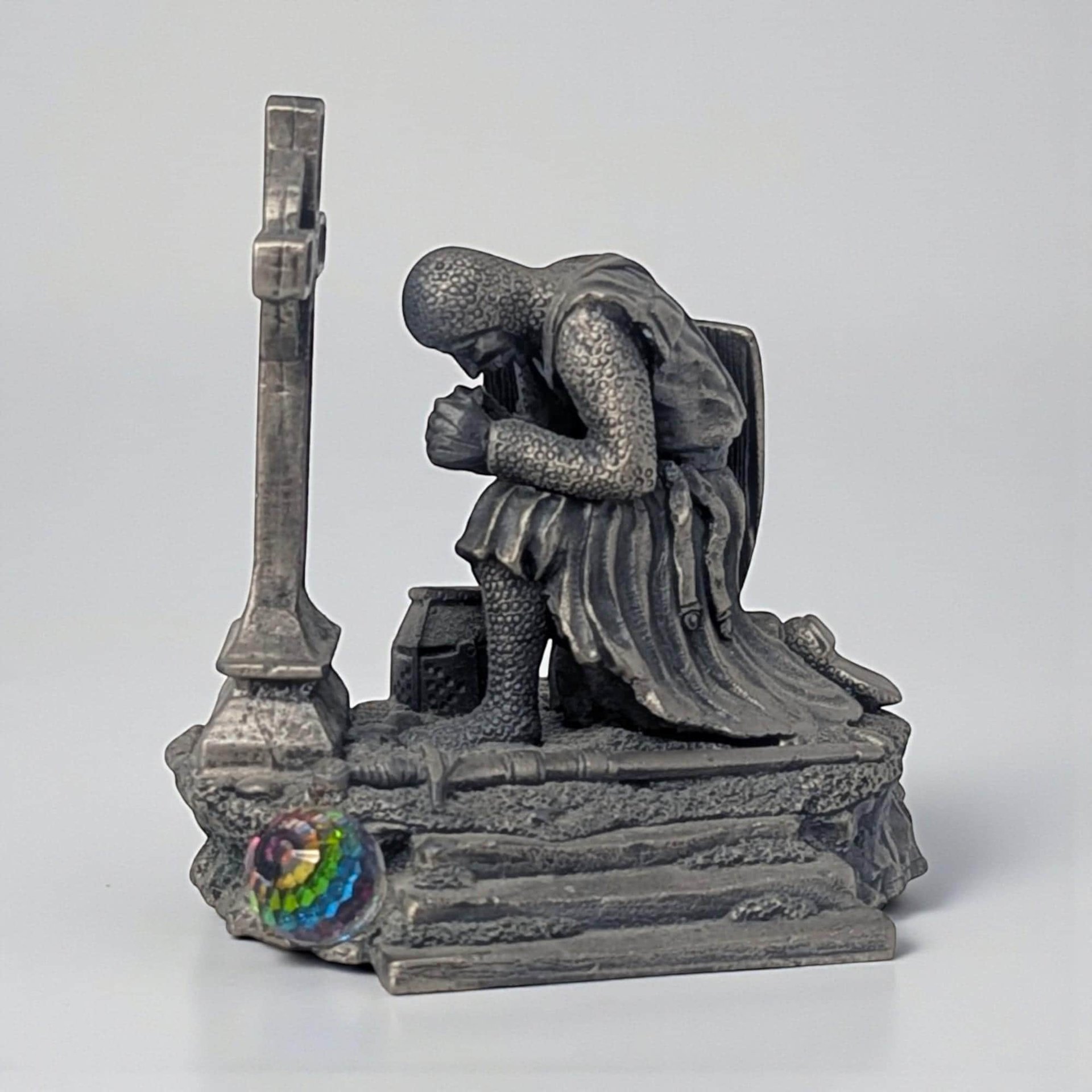 A solid pewter figurine depicting a knight in full armor kneeling in prayer. He rests his hands on the hilt of a sword planted in the ground before him, featuring a soft matte grey finish with intricate chainmail and plate armor textures.