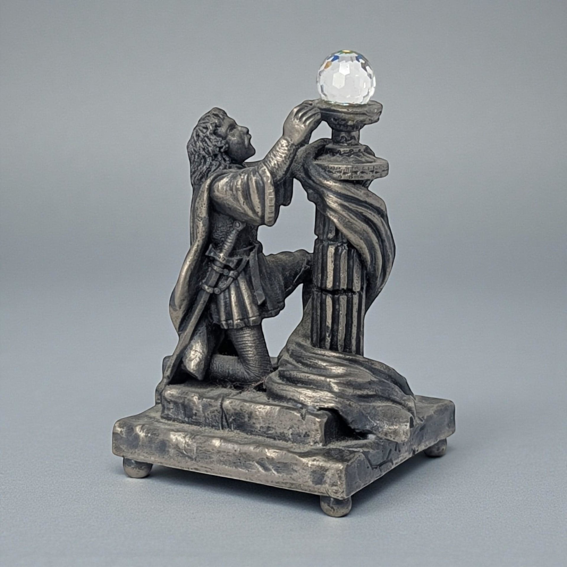 A solid pewter figurine featuring a medieval knight in full plate armor, standing and holding a chalice with both hands. The knight has a detailed surcoat and helmet, with the entire sculpture finished in a soft matte grey antiqued patina.