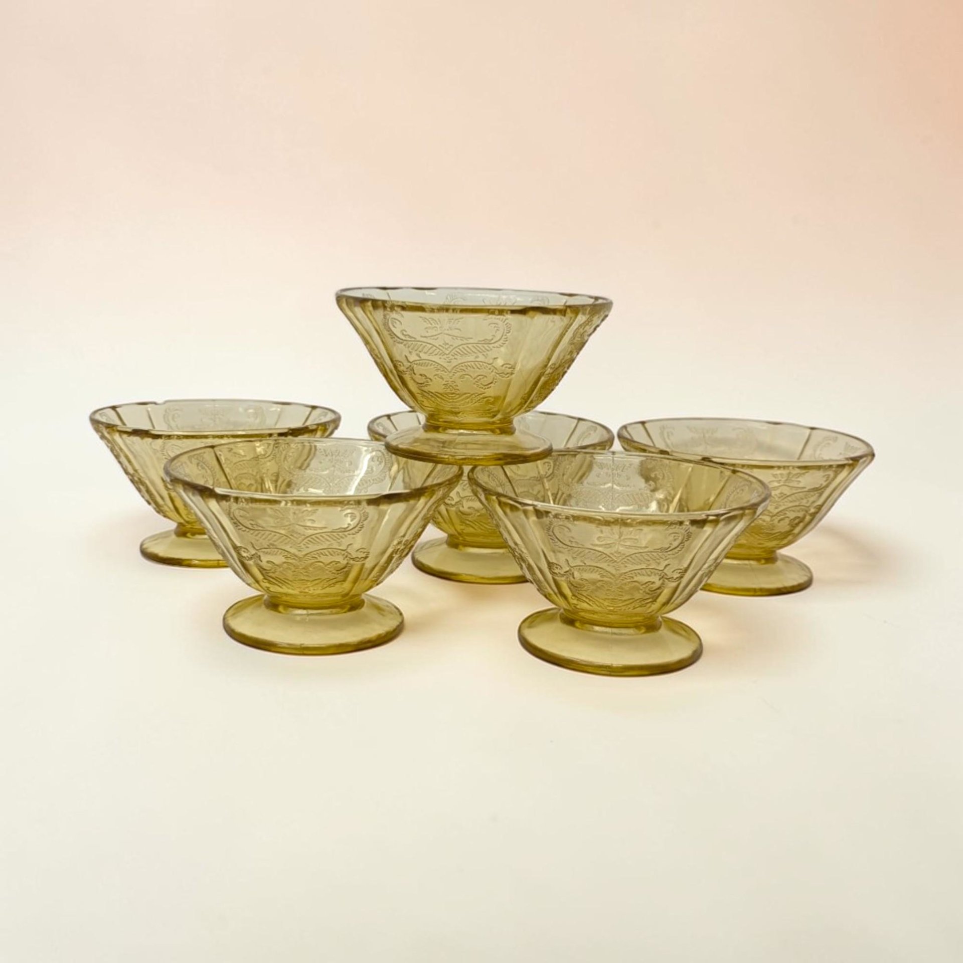 A set of six footed amber glass dessert cups. Each bowl features a flared rim with a lacy, molded scroll and floral pattern. The stems are short and sturdy, leading down to a wide, flat circular base.