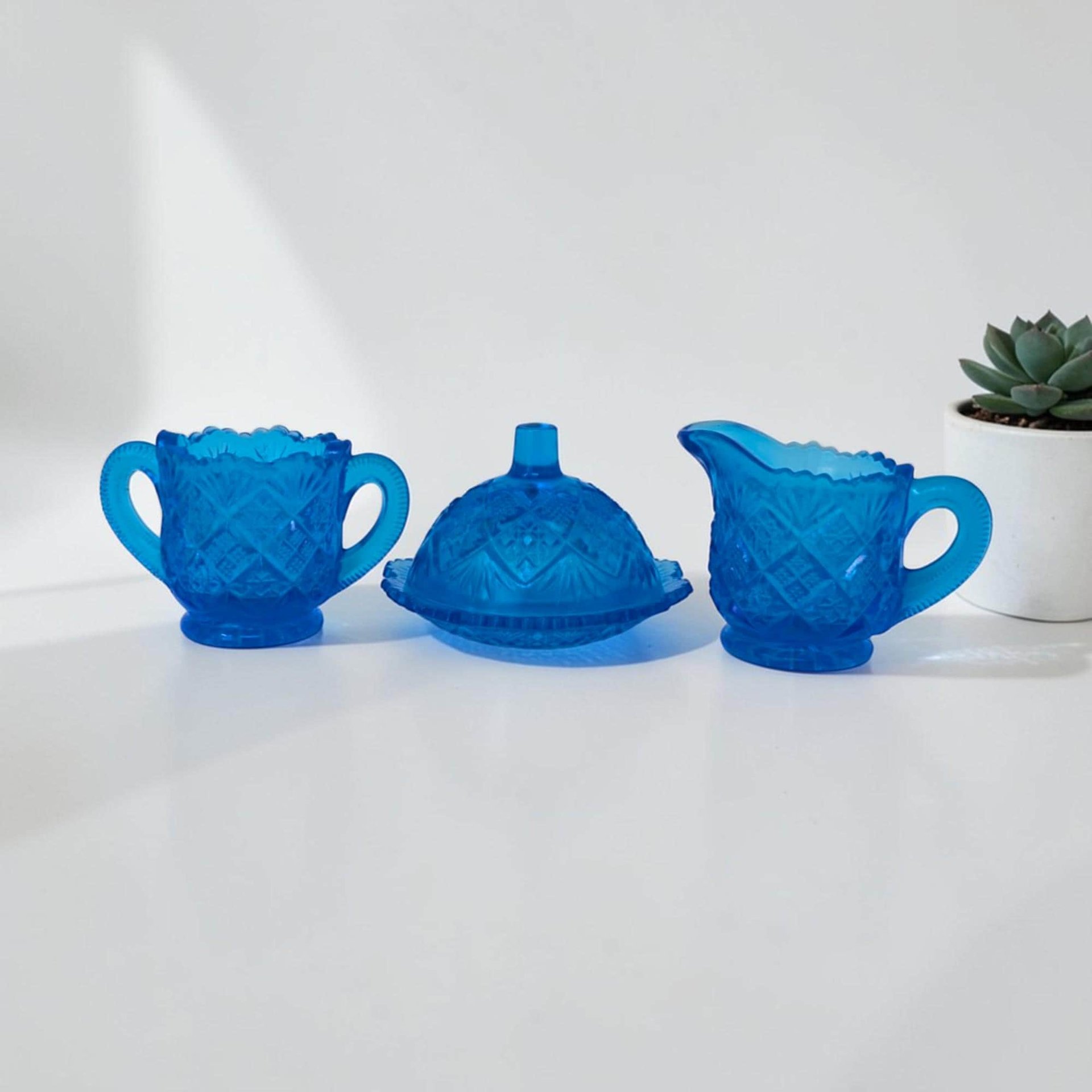 A three-piece miniature toy glassware set in vibrant sky blue. Includes a tiny lidded butter dish, a small creamer, and a matching sugar bowl, all featuring a repeating thumbprint indentation pattern.