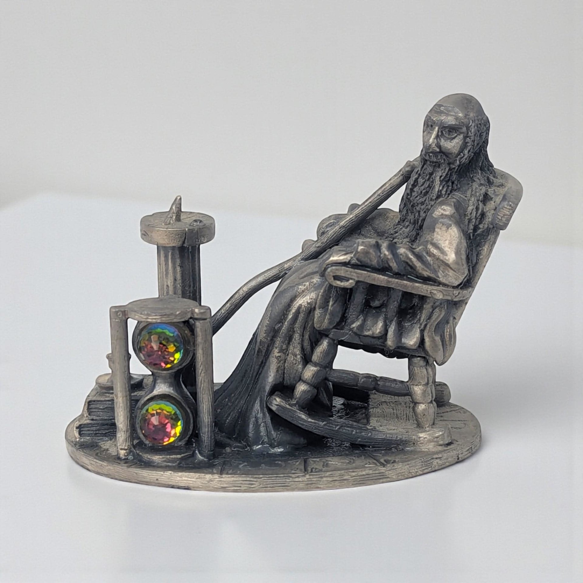 A solid pewter figurine of an elderly bearded man with a long pipe sitting in a rocking chair. A large hourglass stands beside him, containing two iridescent rainbow crystal orbs. The piece has a dark metallic patina and detailed textures.
