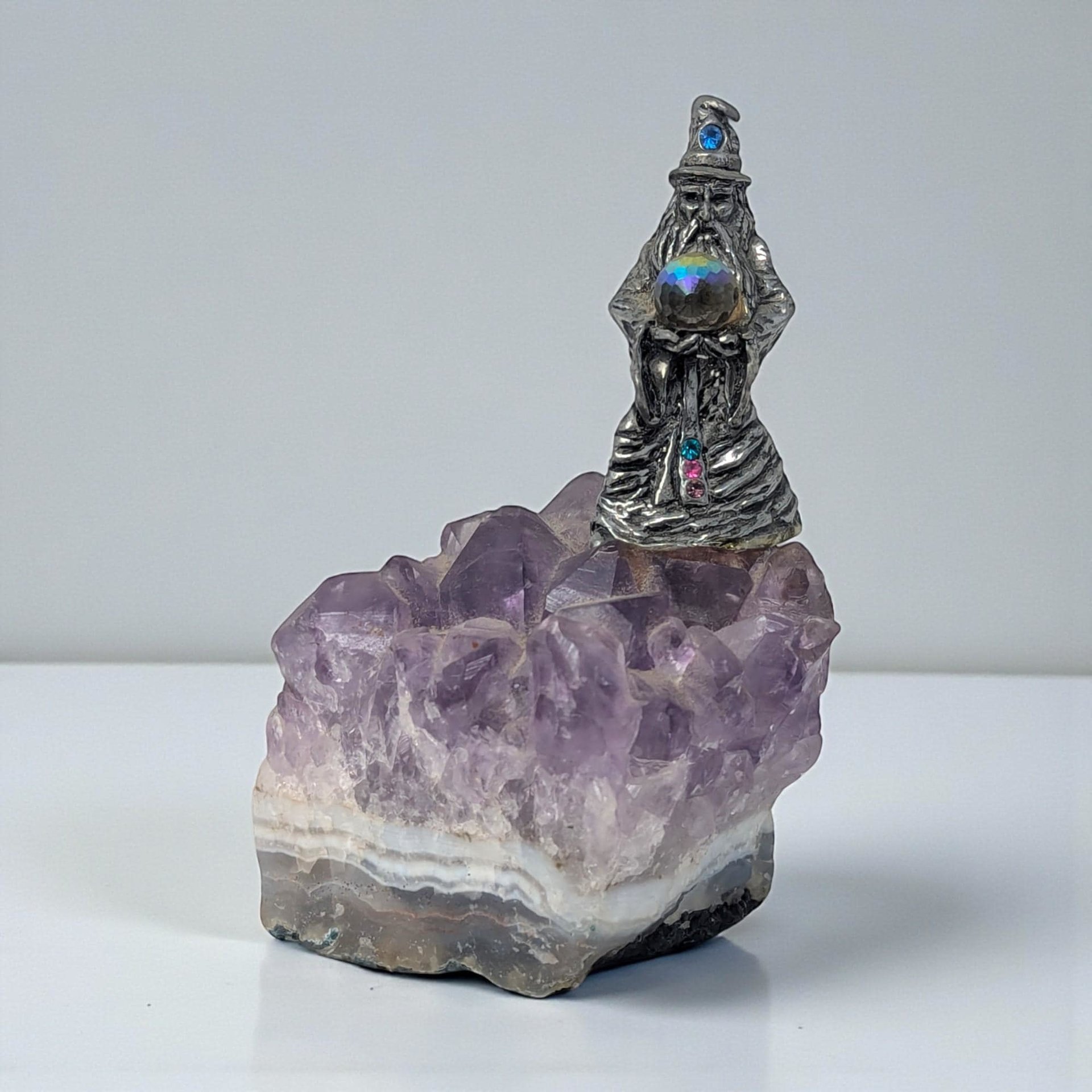 A detailed pewter figurine mounted on a jagged, purple amethyst crystal cluster. The silvery metal features a dark patina in its recessed details, contrasting with the translucent violet hues of the stone base.