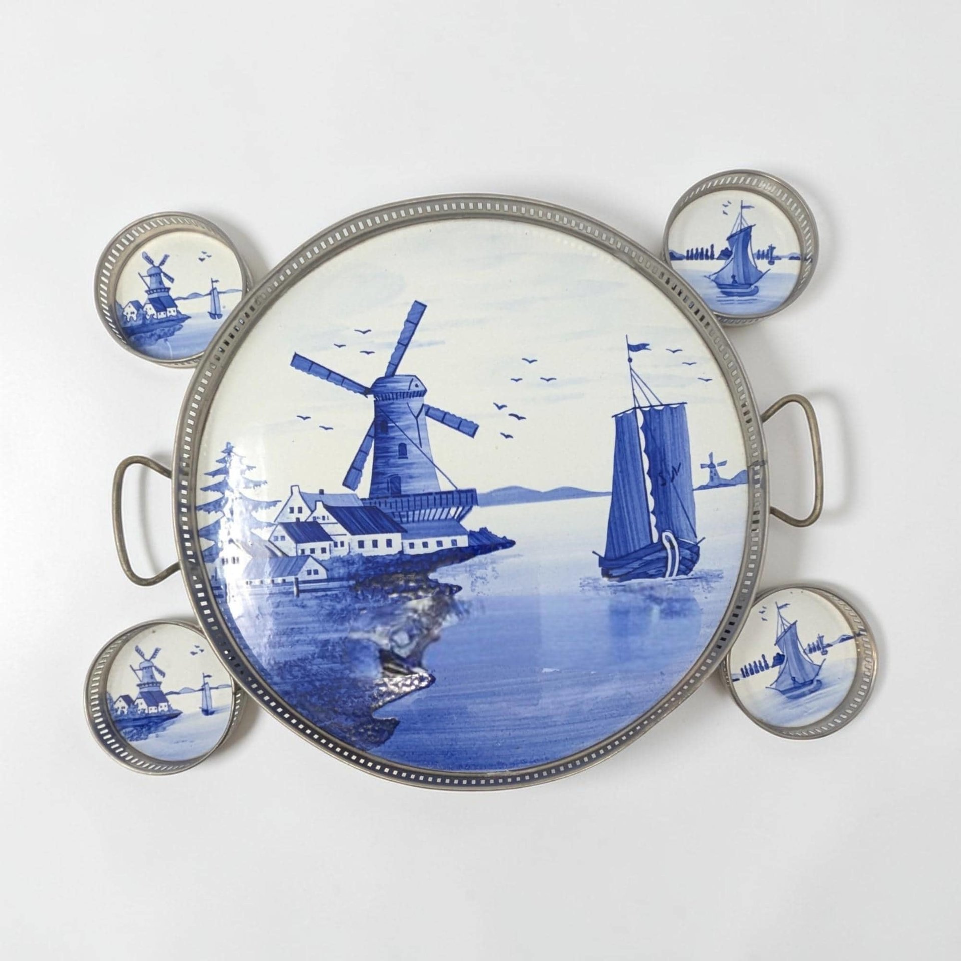 Circular porcelain tray and four matching coasters featuring hand-painted blue windmills and sailboats. The pieces are encased in silver-toned galleried metal rims with a pierced vertical pattern. Smooth glazed finish with aged metal patina.