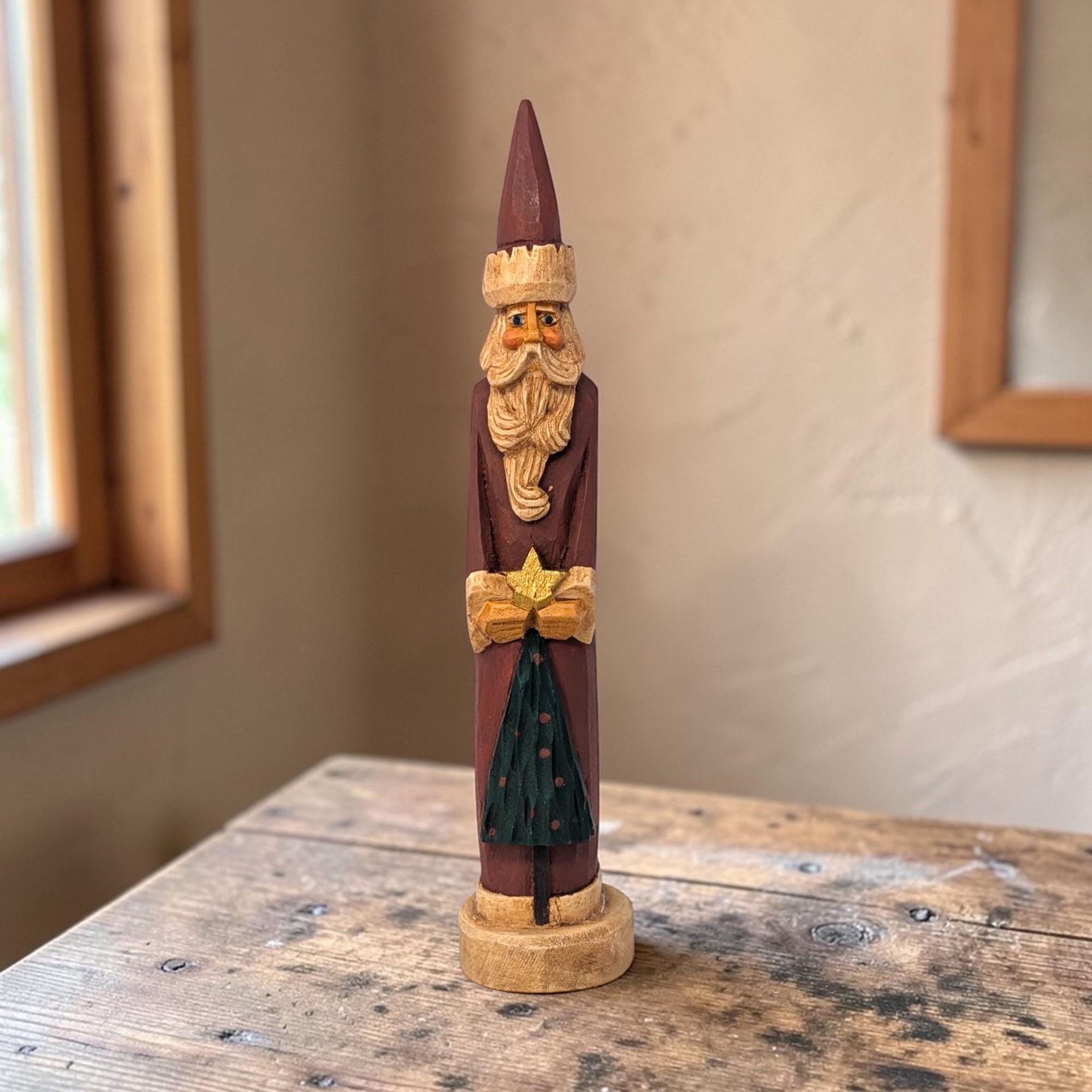 Tall slender hand-carved wooden Santa Claus figurine in a traditional red robe with white trim. The figure holds a small green tree. Natural wood grain visible through the matte paint.