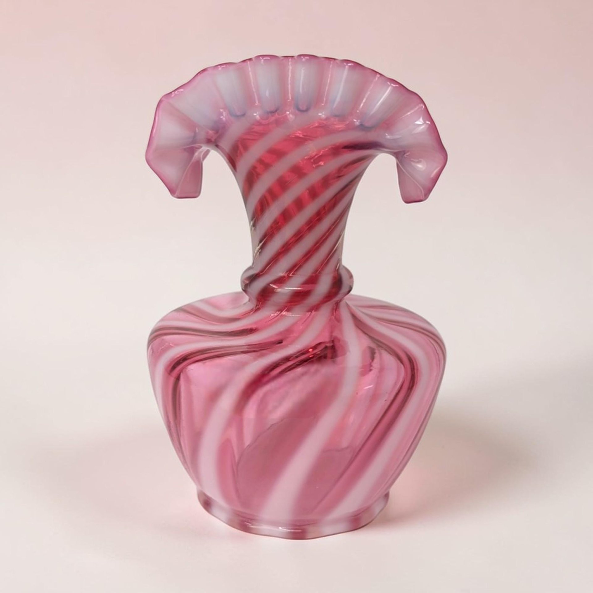 Cranberry pink glass vase with diagonal opaque white opalescent swirls. The silhouette features a bulbous body, narrow neck, and a wide, ruffled fan-shaped rim. The texture appears smooth and glossy with a delicate ribbed pattern.