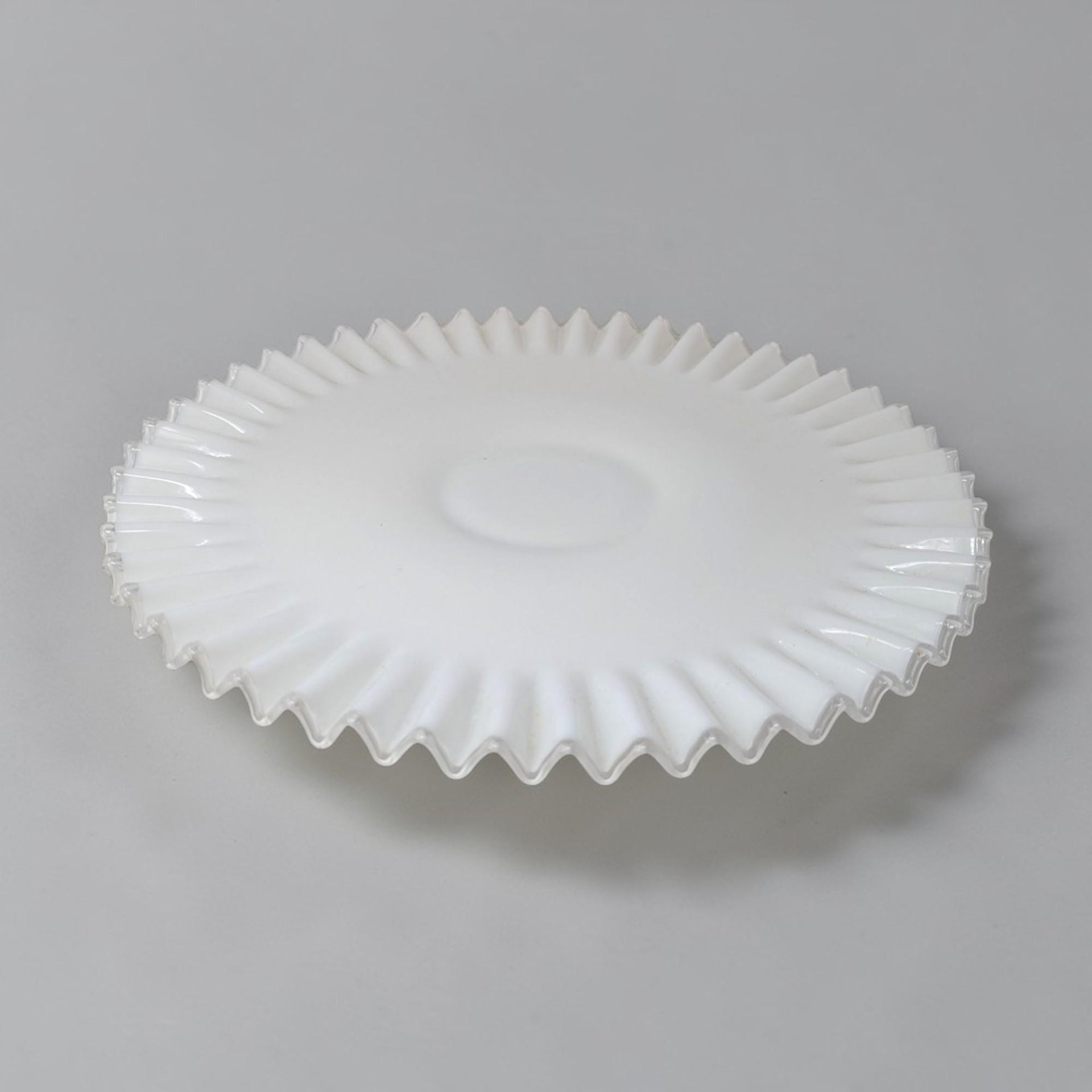 Opaque white milk glass plate with a smooth center and wide, deeply ruffled border edged in a thin ribbon of clear glass. The silhouette is low and circular with a repetitive, undulating texture along the perimeter.