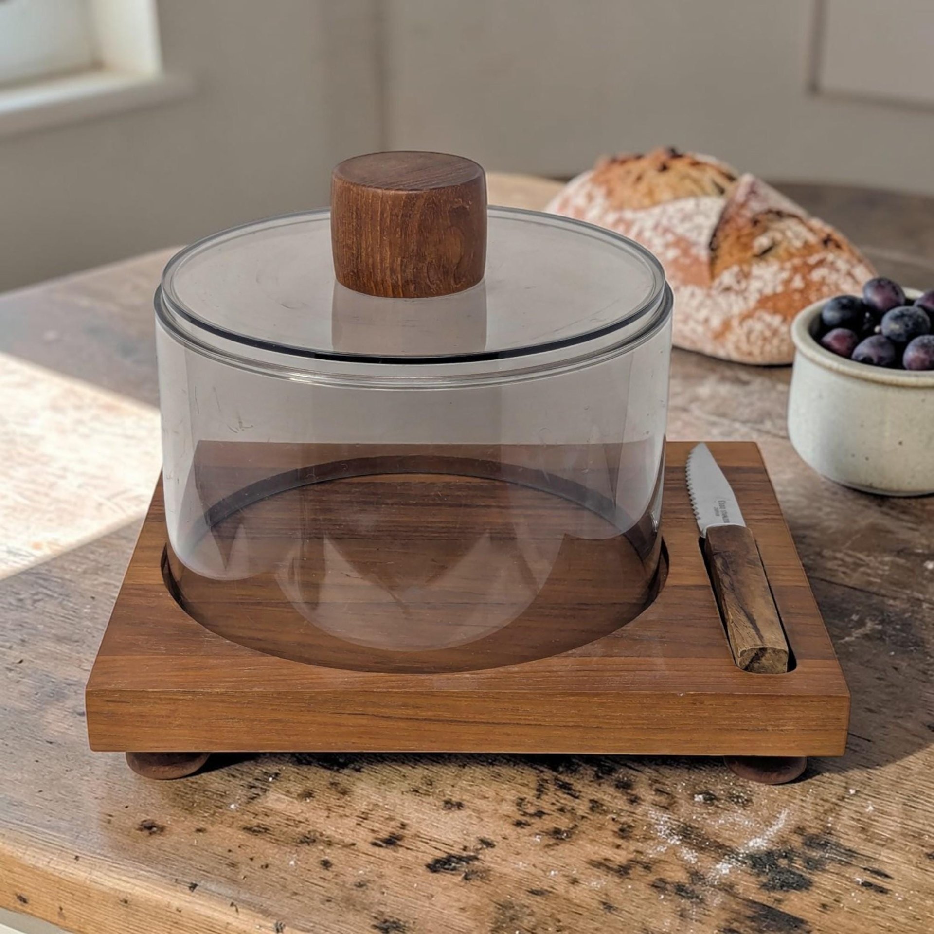 A rectangular teak wood cheese board with a circular recessed center. A cylindrical smoked plastic dome with a round teak knob sits in the groove. A small serrated knife with a matching teak handle rests in a side slot.