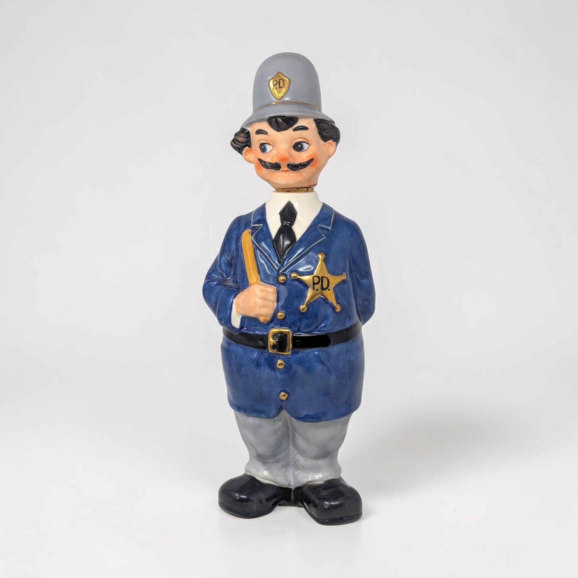 A painted ceramic policeman figurine in a blue uniform and grey helmet with gold "P.D." accents. He has a black mustache, holds a nightstick, and wears a gold star badge. The figure stands on black shoes with hand behind his back.