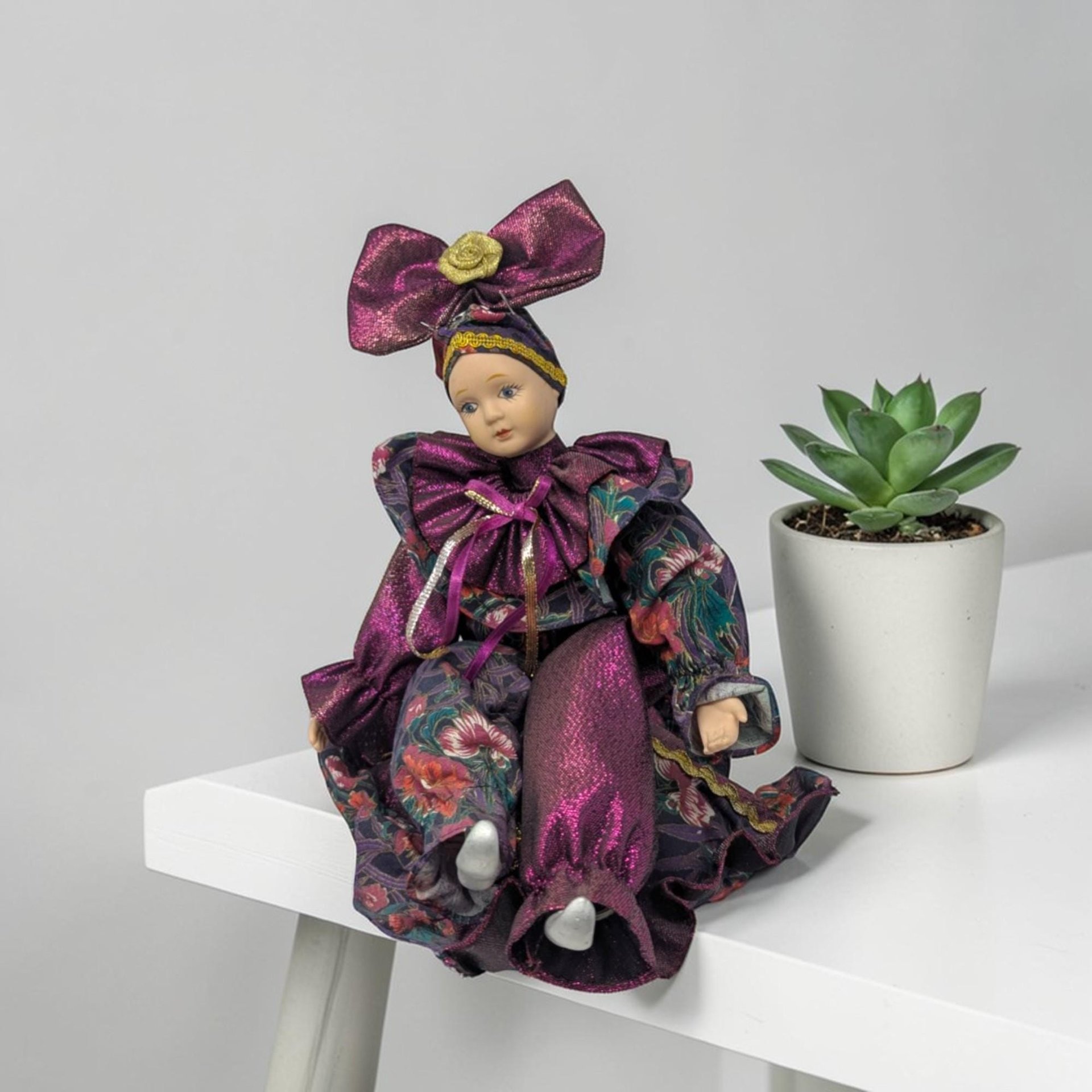A porcelain harlequin doll wearing a shimmering magenta and floral patterned jester costume. The doll features a large magenta bow with a gold rose center on its head and silver-painted shoes. It sits in a relaxed pose with delicate facial features.