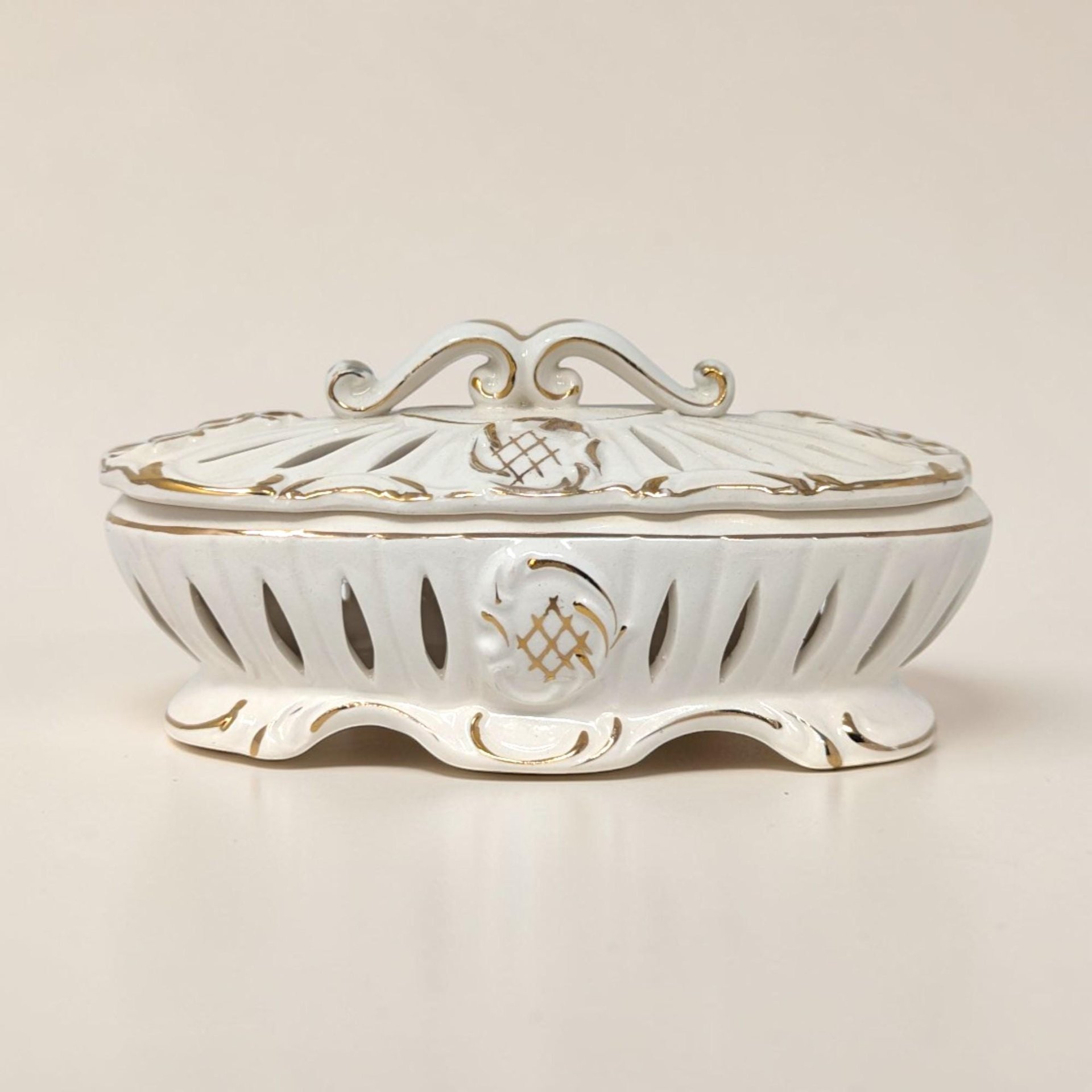 An oval ivory ceramic trinket dish with a matching lid. Both pieces feature vertical reticulated cutouts and gold-painted scroll edges. The lid has a decorative scrolled handle, and the body sits on a scalloped pedestal base with gold accents.
