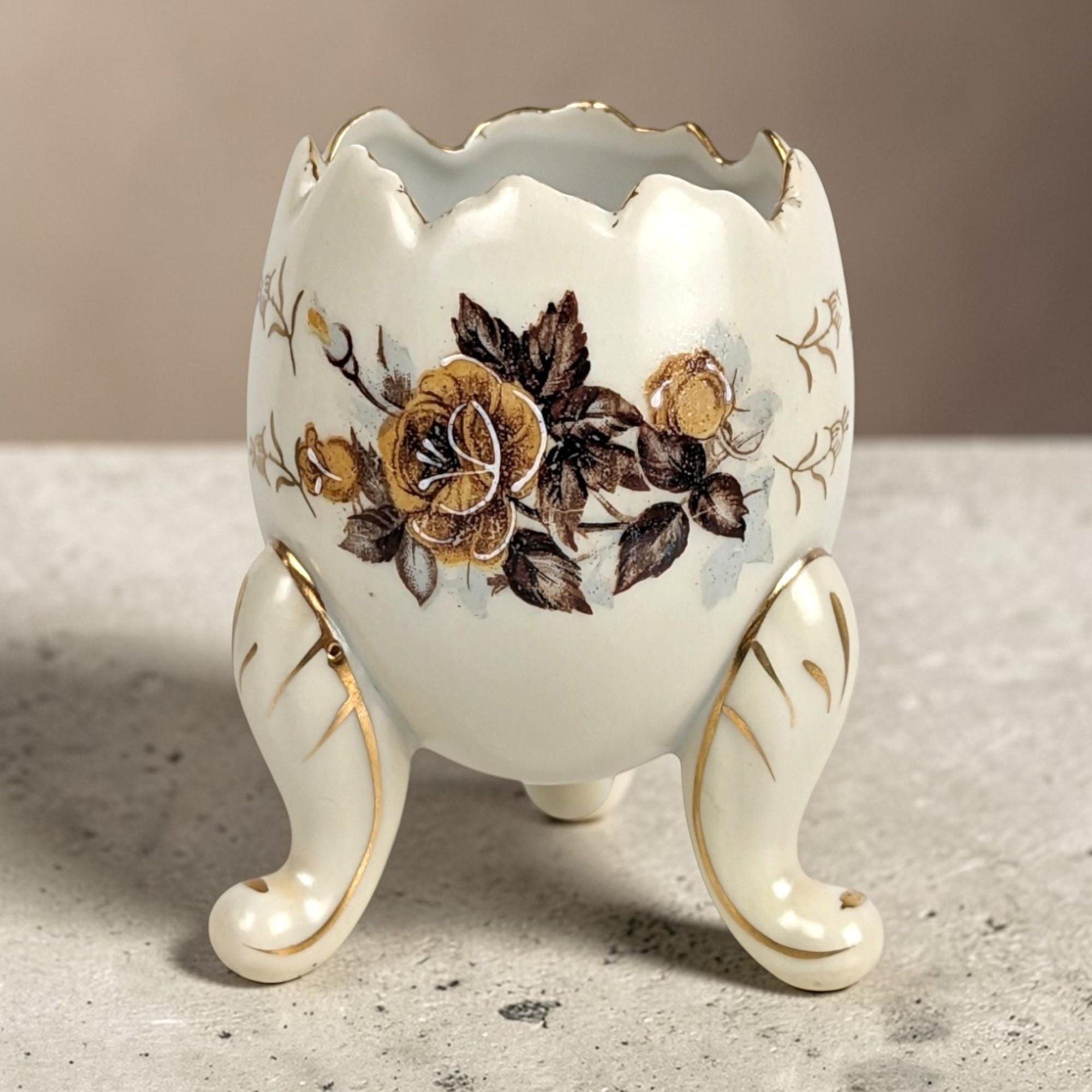 Cream porcelain egg-shaped vase with a jagged rim and three gilded scroll feet. Features a central transfer of golden-brown roses and smaller gold leaf sprigs. Smooth, glazed ceramic texture with gold trim on the feet and scalloped edge.