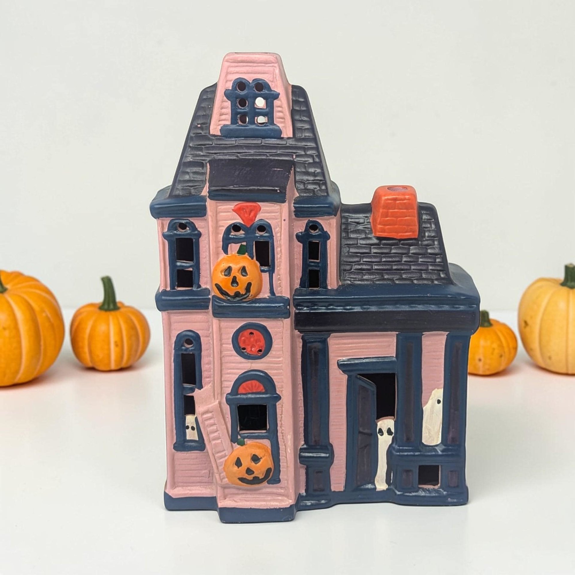 A hand-painted ceramic haunted house with pink siding and deep navy blue trim and roofing. The Victorian silhouette features orange jack-o'-lanterns on the balcony and porch, white ghosts peeking from windows and doorways, and a bright orange chimney.