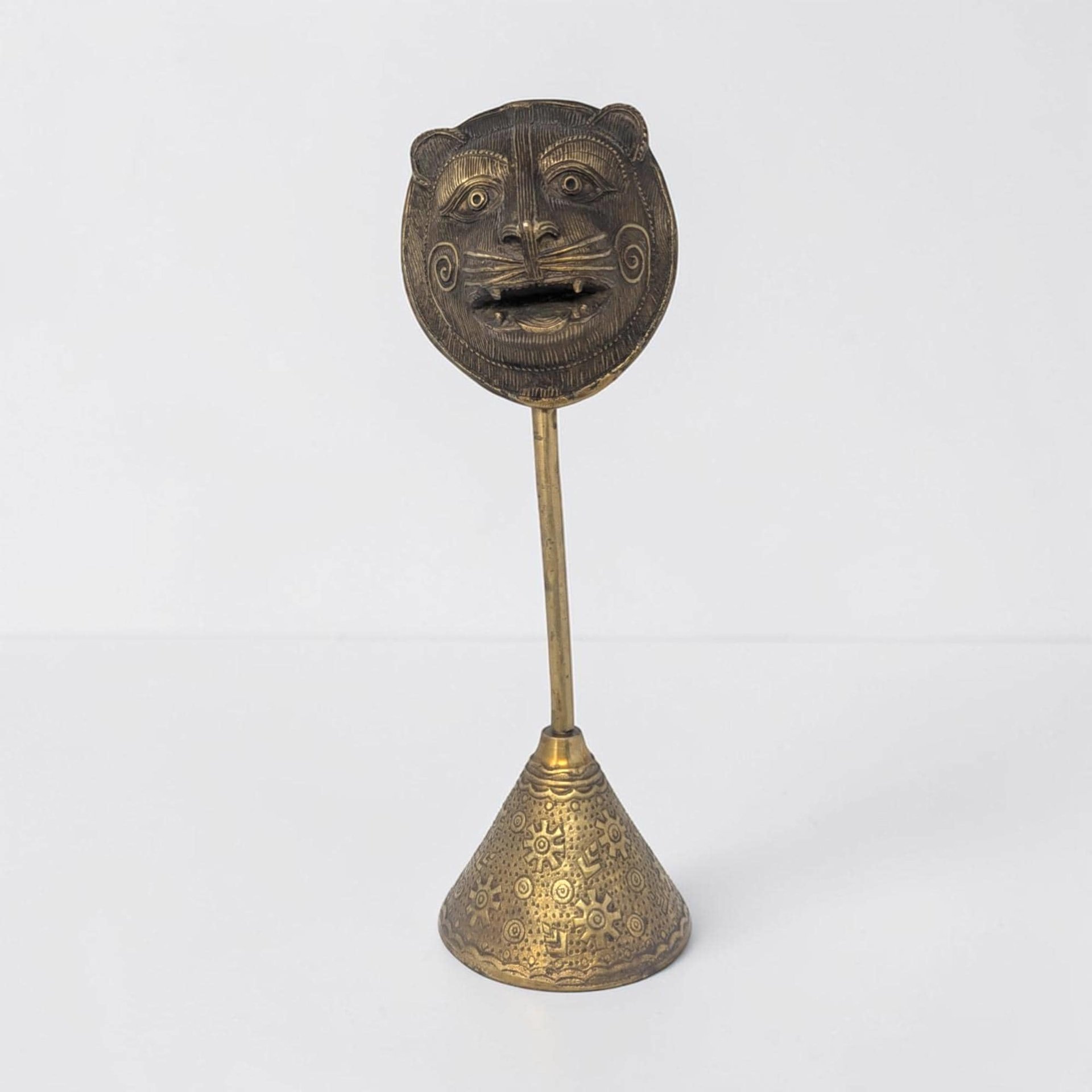 A tall brass sculpture featuring a stylized tiger or lion face with large eyes and an open mouth. The mask is mounted on a slender rod fixed to a conical base decorated with sunburst and geometric engravings.