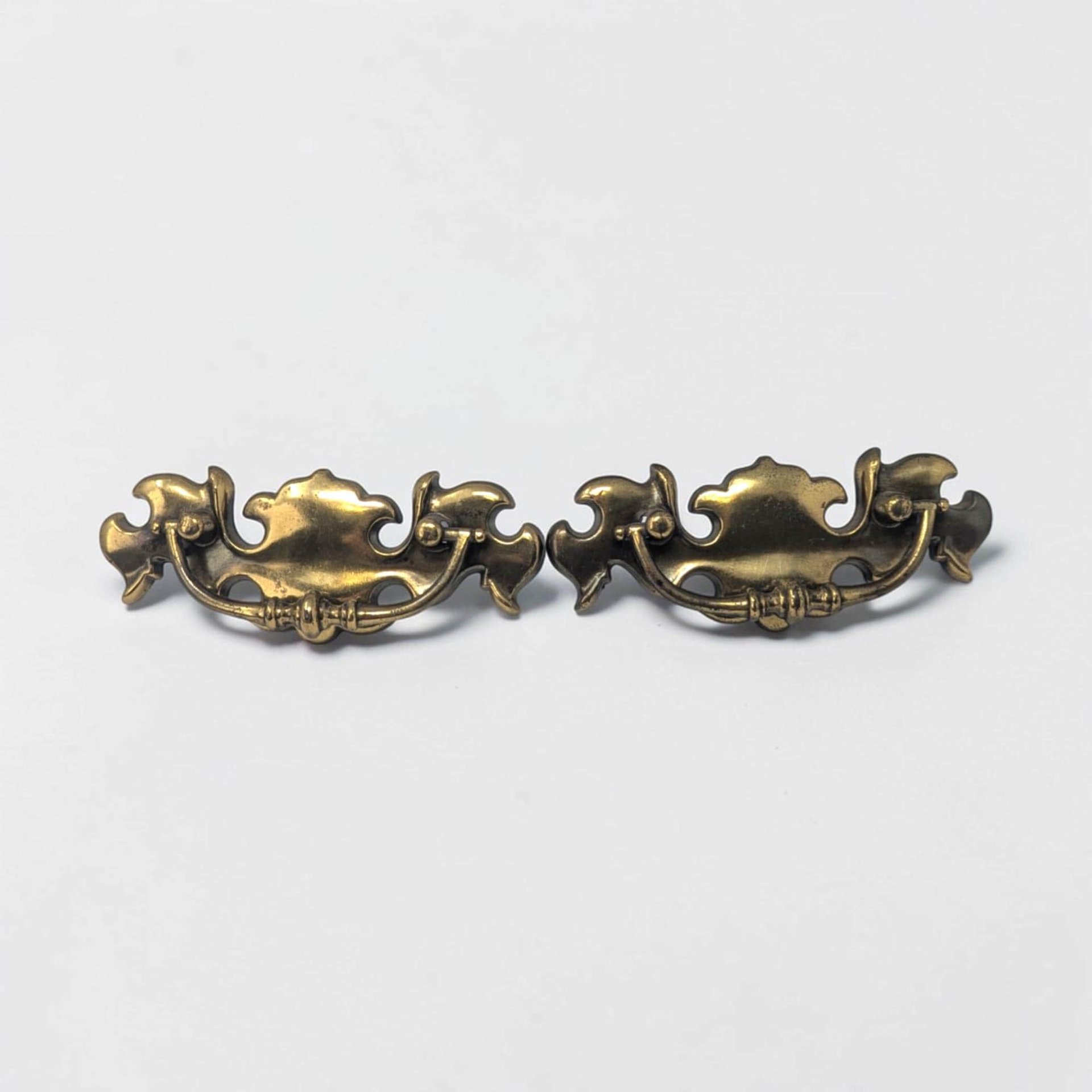 A pair of brass drawer pulls featuring an ornate batwing-shaped backplate and a curved bail handle. The metal has a deep, aged gold patina with visible swirling flourishes and traditional mid-century decorative etching.