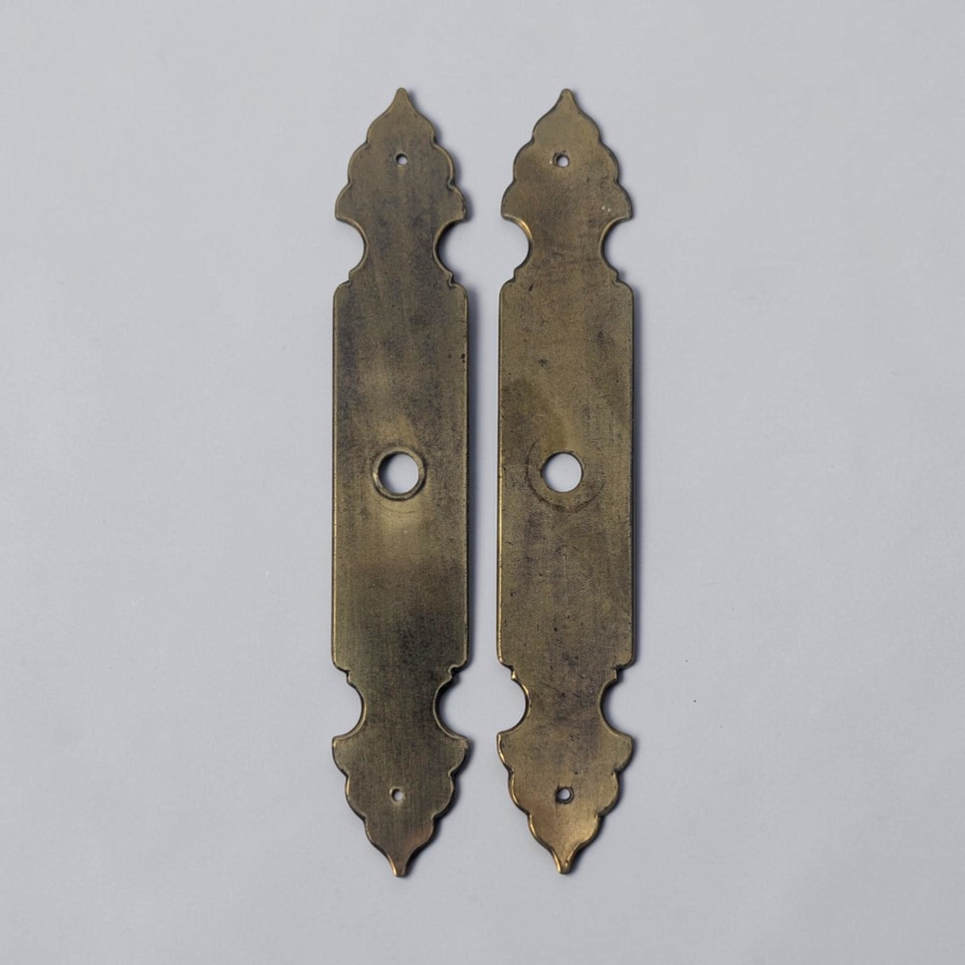 A pair of long brass escutcheon plates with a rectangular center and ornate scalloped ends. The metal has a heavily aged, dark gold patina with central circular cutouts and smaller mounting holes at each tip.