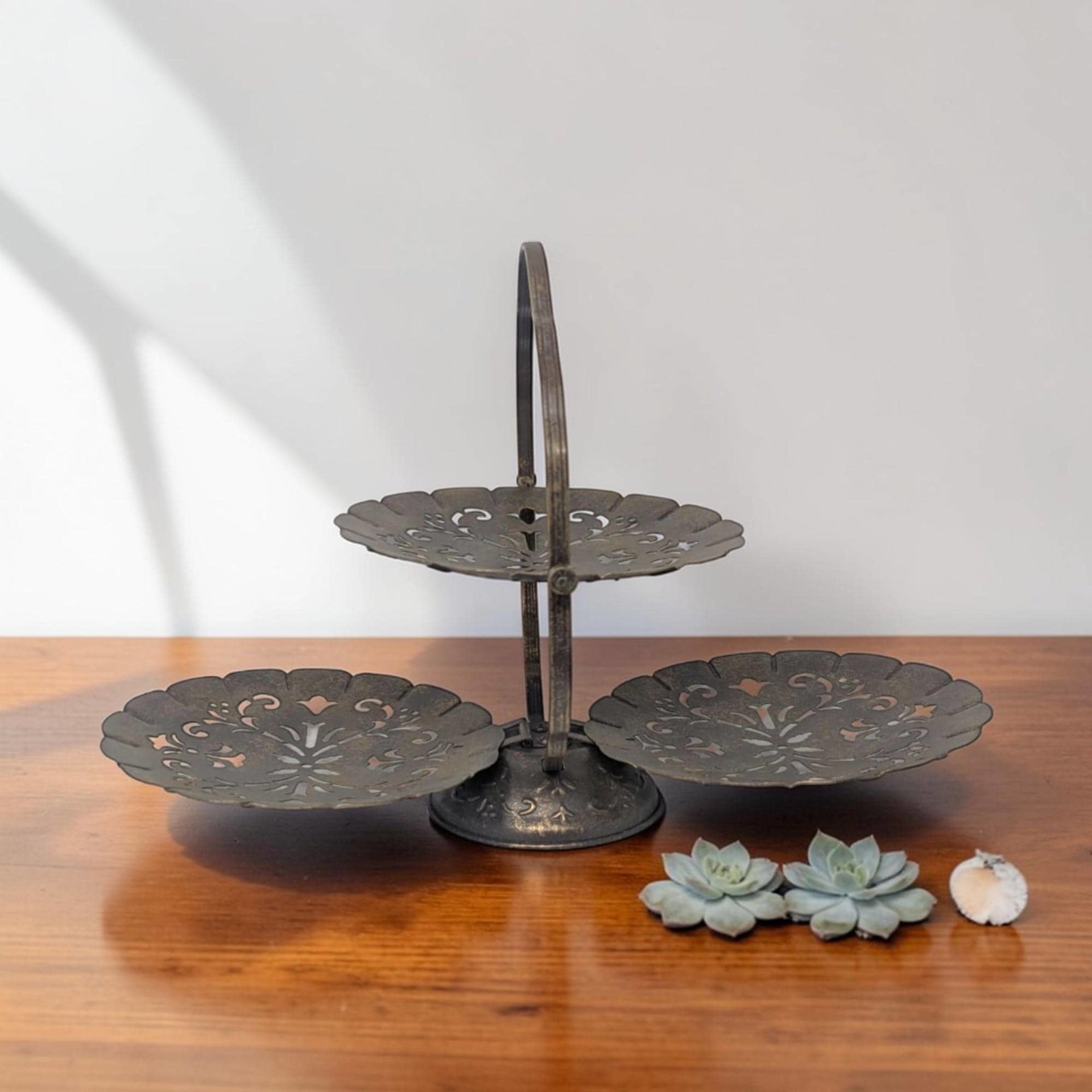 A vintage three-tiered metal serving tray with a folding circular handle and scalloped filigree plates. The dark, aged metal features intricate cutout patterns on each tier, supported by a central vertical rod and a weighted decorative base.