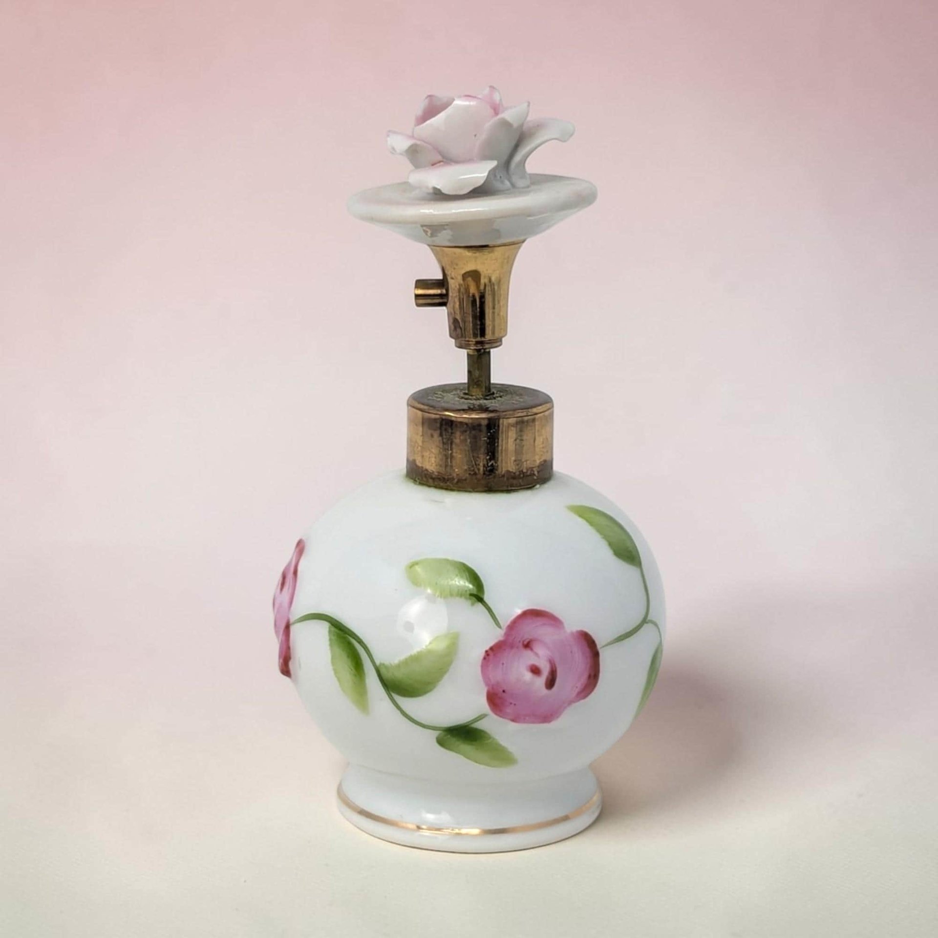 A 3.75-inch bone china perfume bottle with a round bulbous silhouette and a smooth white porcelain texture. It features hand-painted pink roses with green leaves and a sculptural rosebud stopper on a gold-tone metal stem.