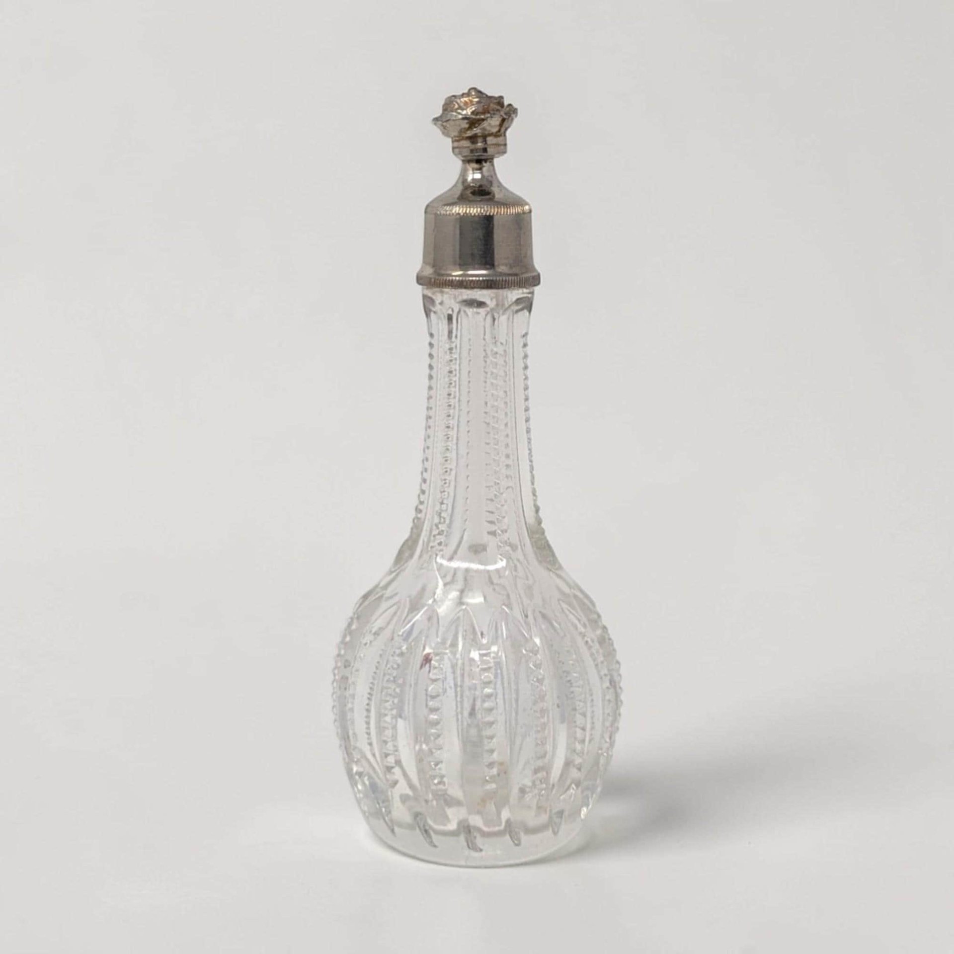 A 5-inch clear glass bottle with a teardrop silhouette and a ribbed faceted texture. It features a long slender neck topped with a decorative silver-tone metal cap featuring a rose floral finial.