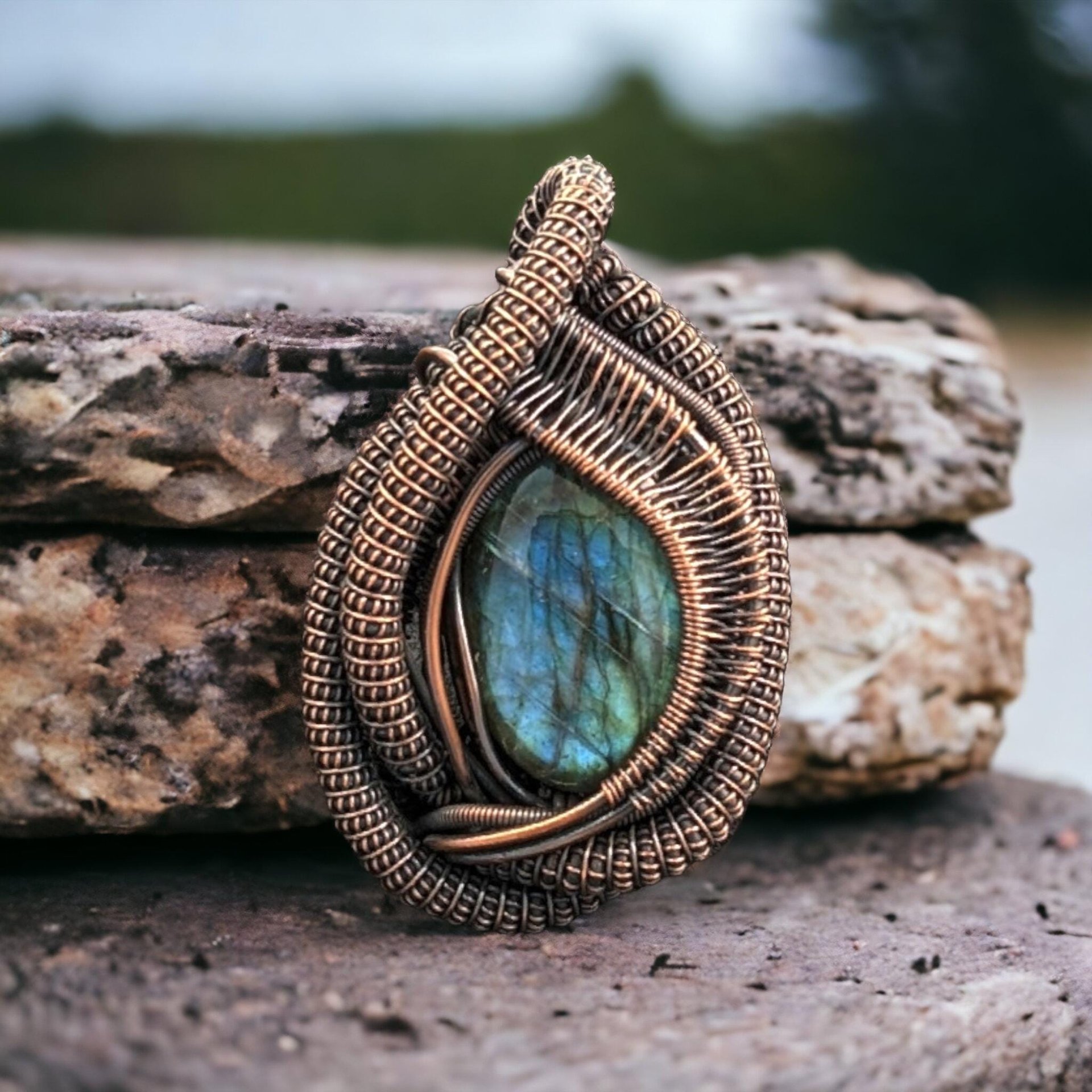 A vibrant blue-flash labradorite cabochon secured in an intricate copper wire weave. The metal features swirling spiral patterns and a textured, oxidized finish against the smooth, dark stone.