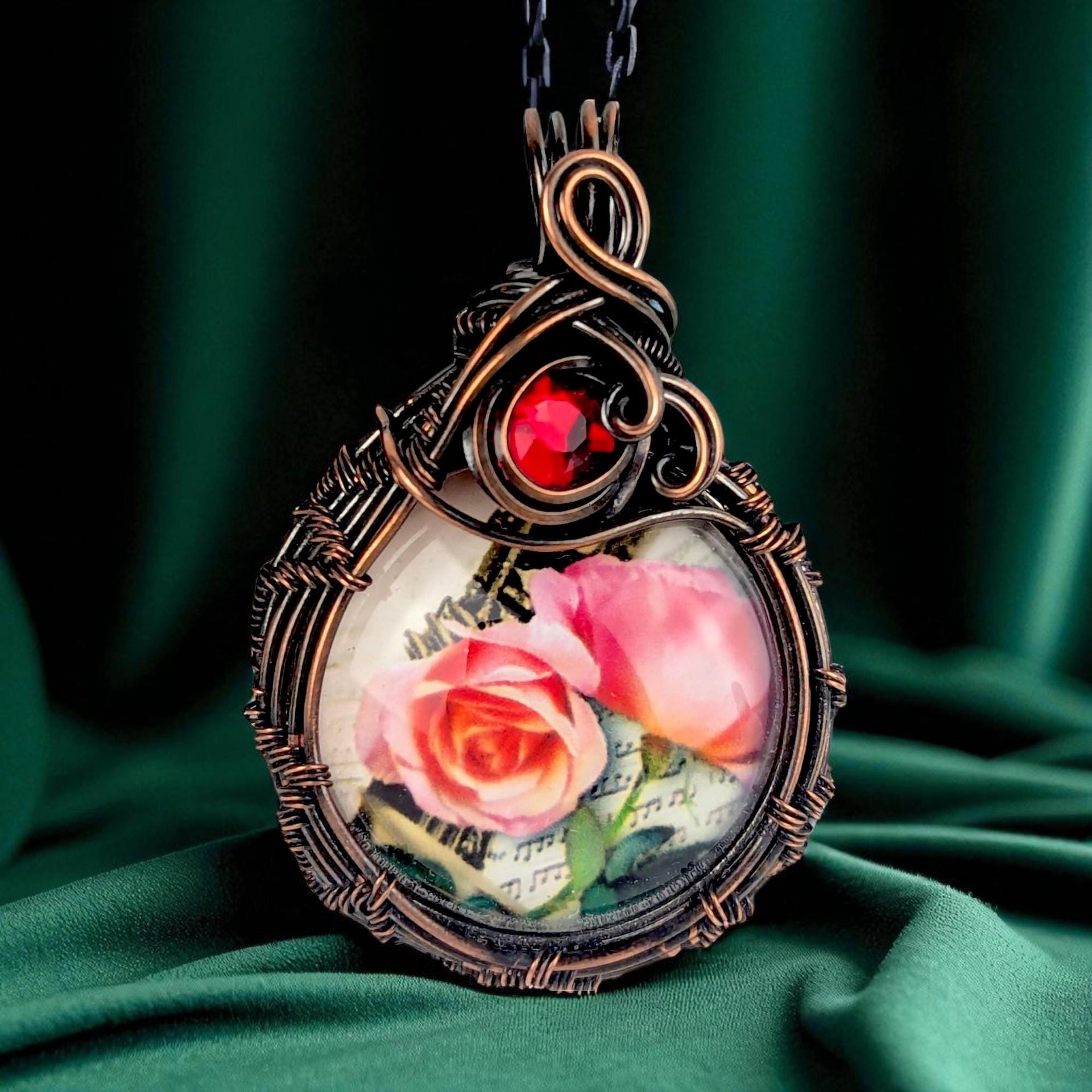 A circular cabochon featuring two blooming pink roses on a white background, encased in complex oxidized copper wire weaving. A single crimson glass bead is set into the ornate wire bail at the top.