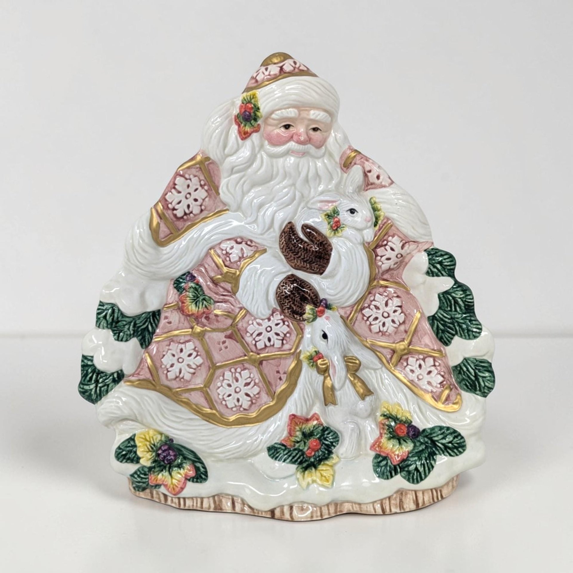Glossy ceramic wall plate shaped like Santa. He wears a dusty rose quilted robe with white snowflake accents and gold trim. He holds two white rabbits. Texture includes raised white fur trim and a base of green boughs and berries.