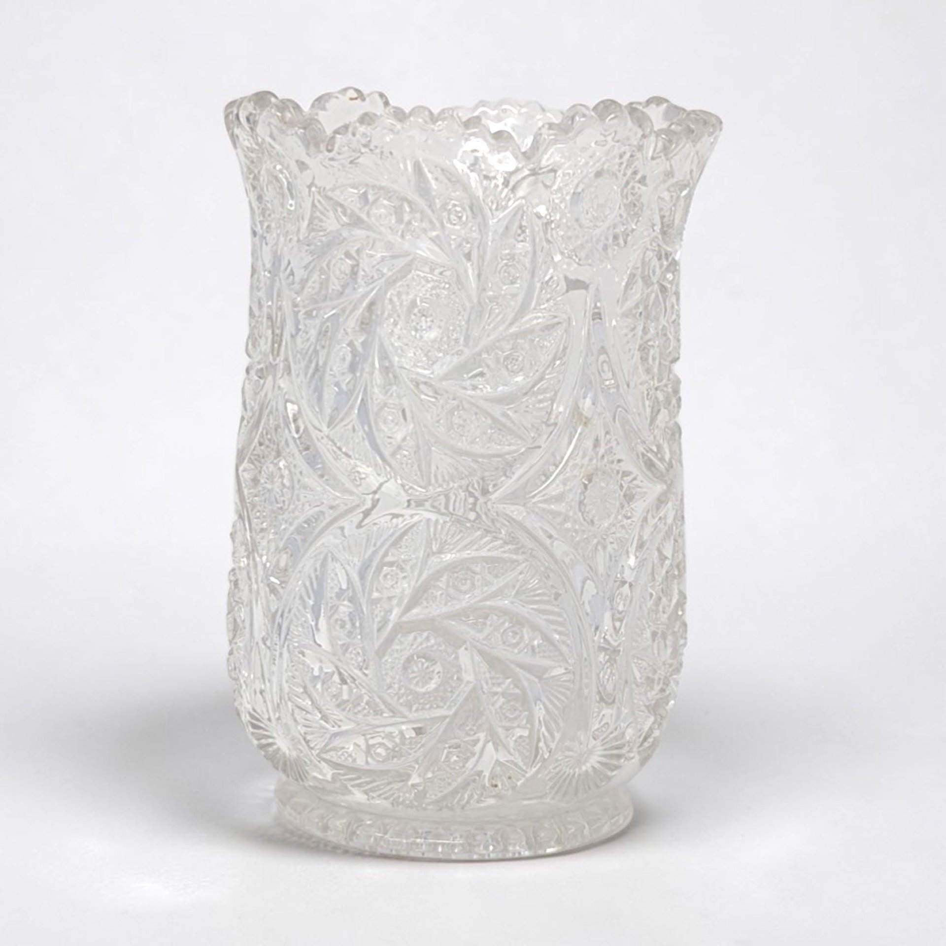 Heavy clear crystal vase with a scalloped rim. Intricate deep-cut pinwheel and starburst patterns cover the entire exterior. Cylindrical form with a slightly flared top and a sturdy pedestal base.