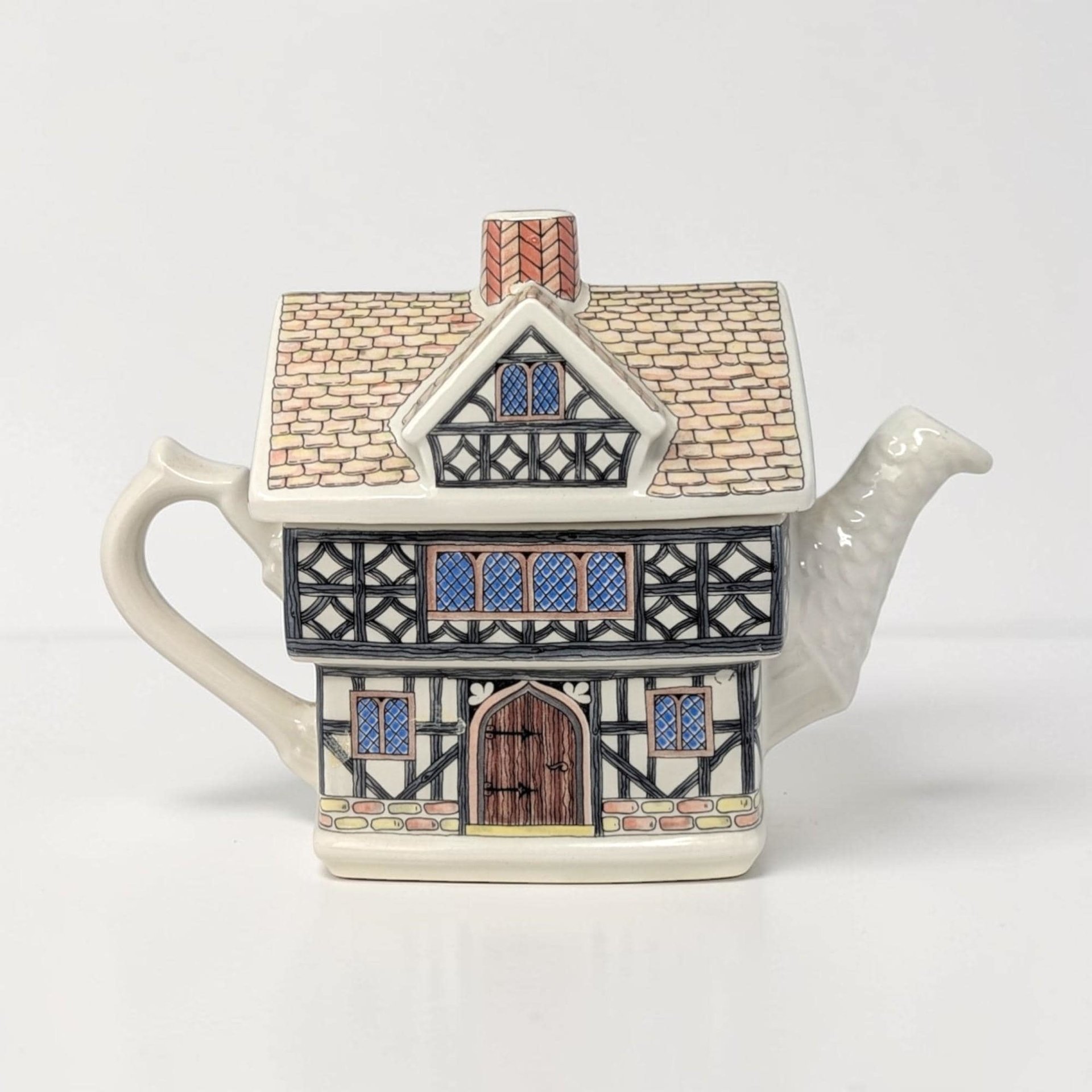 Rectangular ceramic teapot with a high-gloss finish. The cream-colored body features detailed brown and black timber-frame transfers. The lid is a solid brown thatched-roof style with a curved handle and spout.