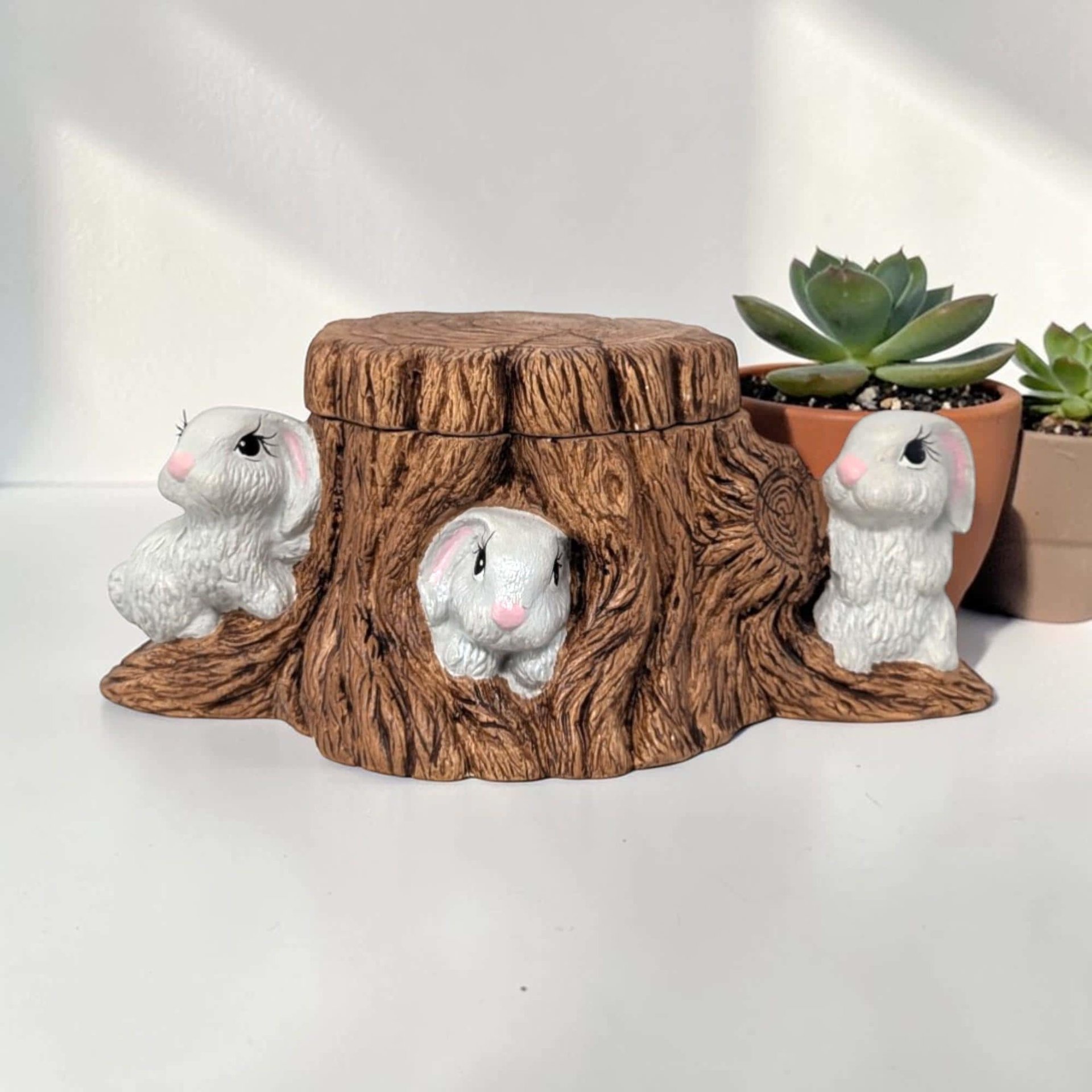 Hand-painted ceramic candy dish shaped like a hollowed-out tree stump. Two white rabbits with pink ears are nestled at the base among green grass. The stump is textured with brown bark and knots.
