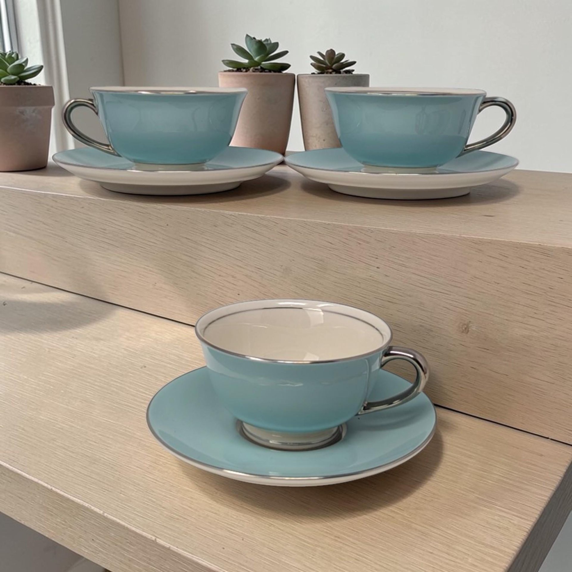 Set of three white fine china tea cups and matching saucers. Each piece features a thin polished silver rim. The cups have a low wide profile with a dainty looped handle and a high gloss finish.