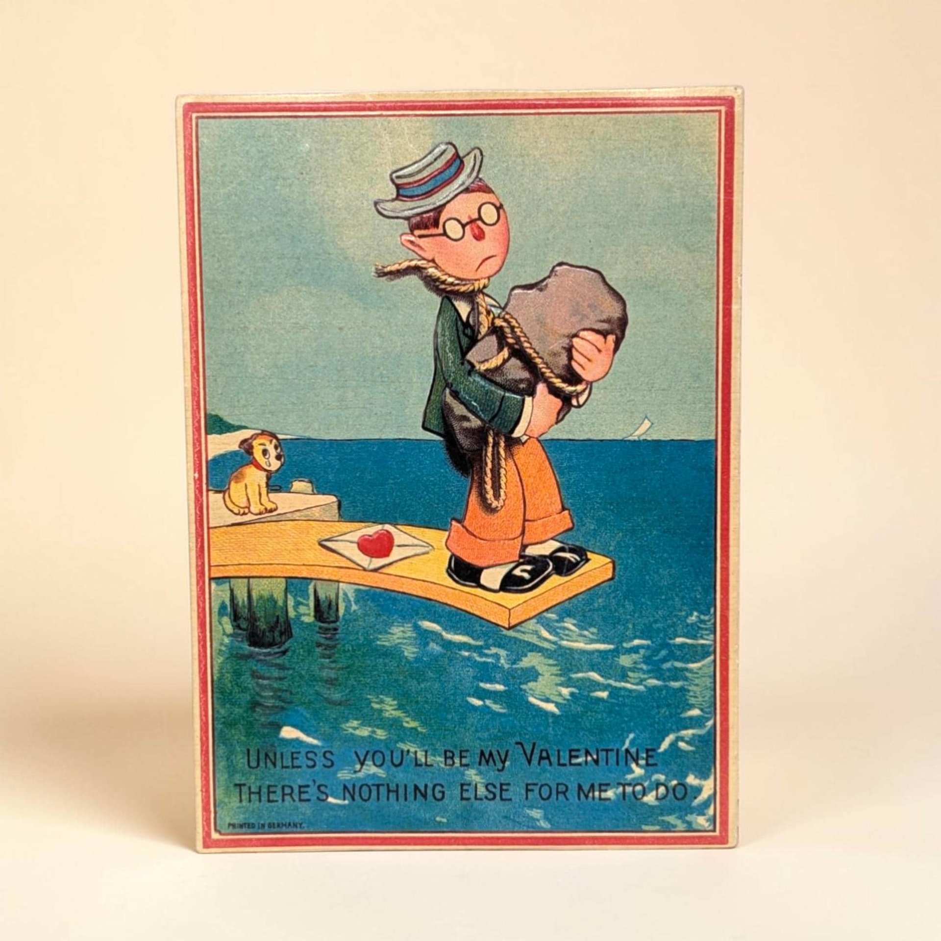 A lithographed postcard featuring a sad man in a straw hat and glasses holding a large stone with a rope around his neck. He stands on a pier above blue water. The colors are muted primary tones with a matte paper texture and red border.