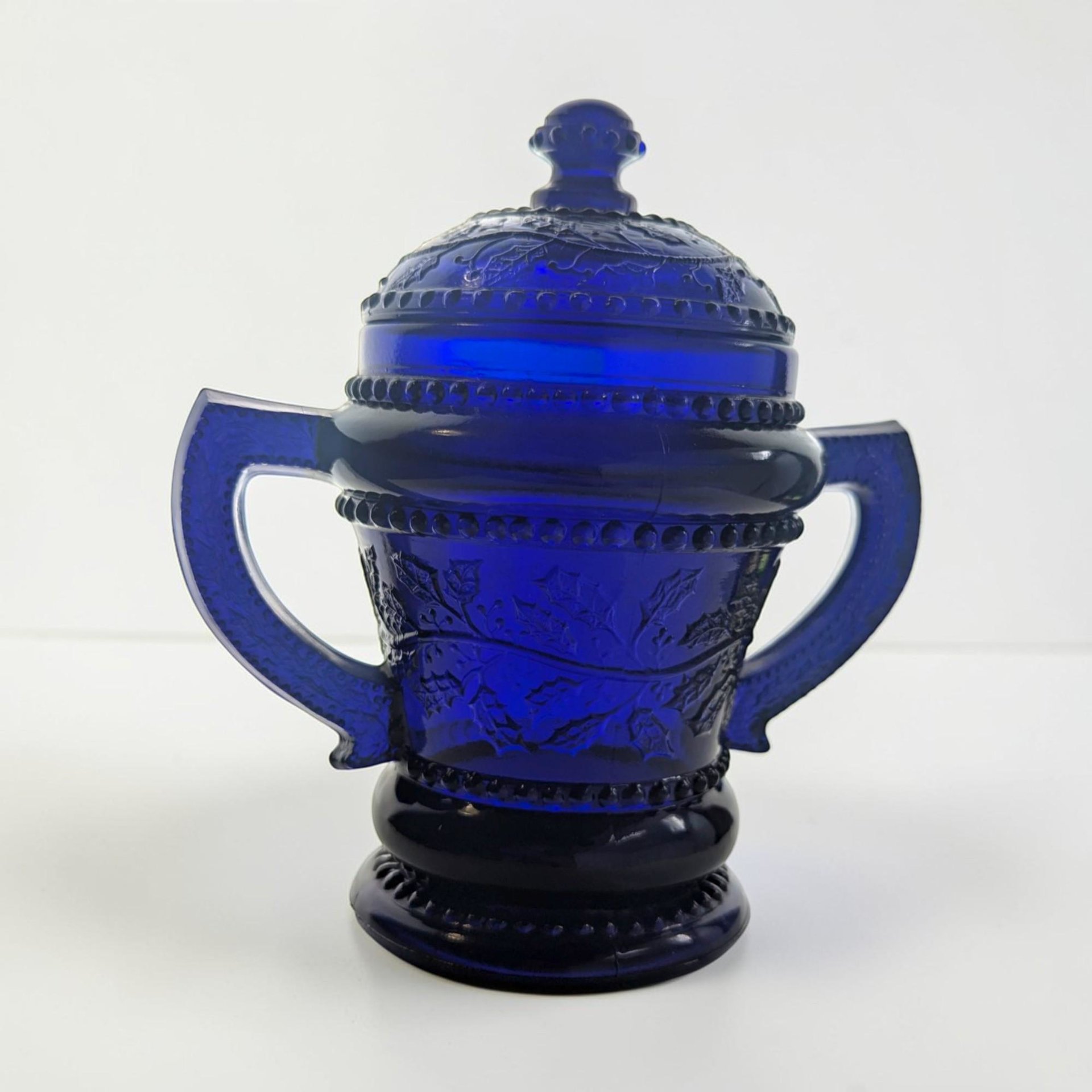 A deep cobalt blue translucent glass sugar bowl with a matching lid. It features two angular side handles, a footed base, and raised patterns of holly leaves and berries surrounded by beaded bands.