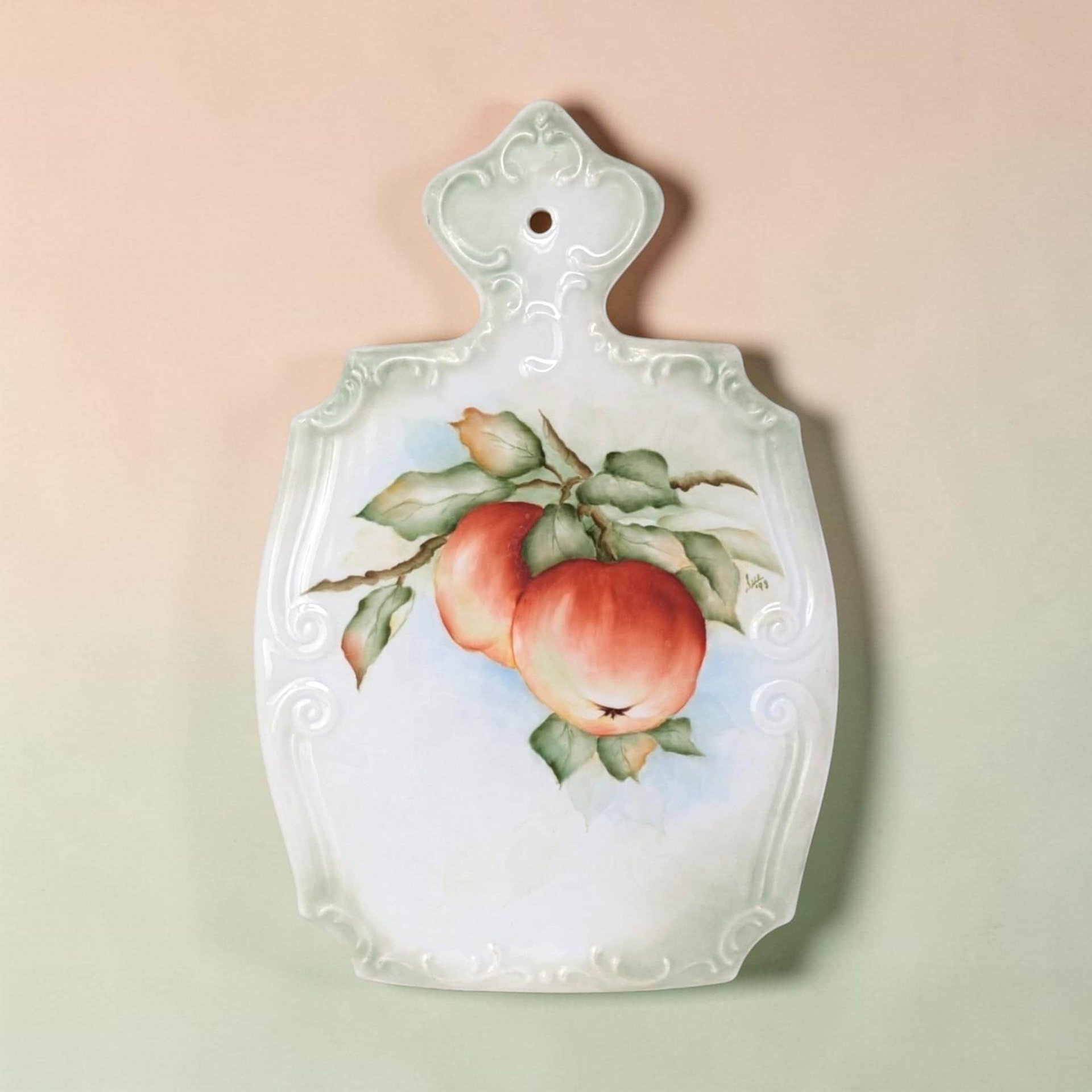A shield-shaped porcelain wall plaque with a decorative scrolled top and scalloped edges. It features a handpainted center illustration of two red apples on a leafy branch against a pale blue and white background with a soft green border.
