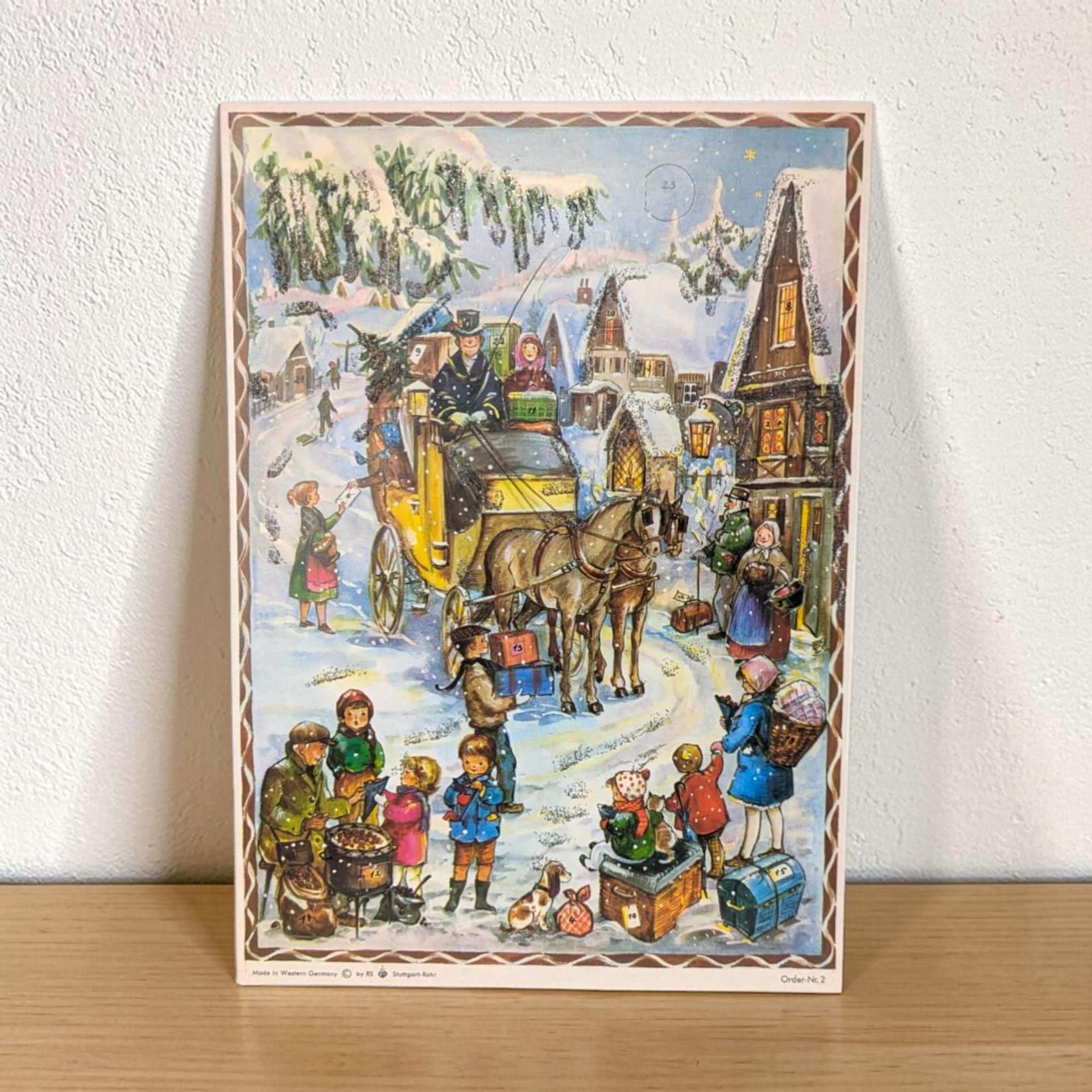 A traditional paper advent calendar featuring a vibrant lithograph of a snow-covered German village at twilight. The rectangular silhouette is accented with delicate iridescent glitter. The numbering on the small perforated doors is clear and crisp.