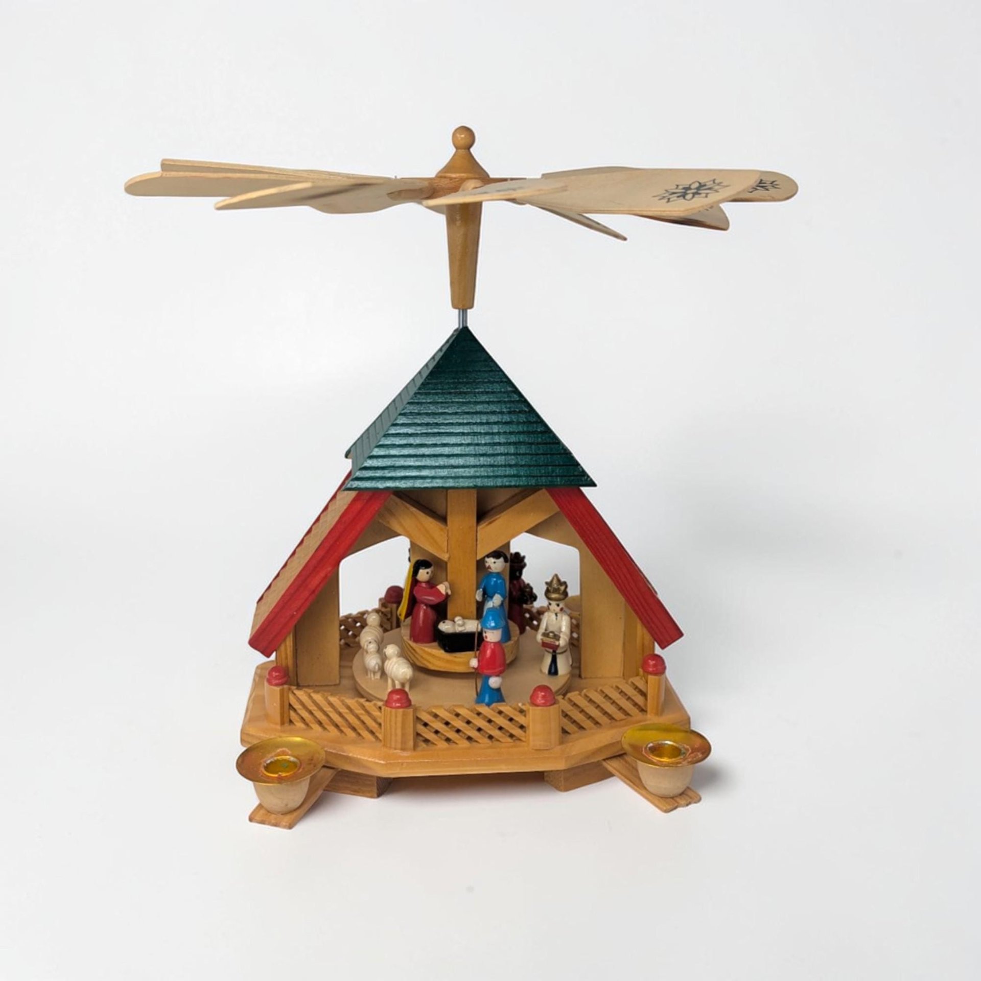 Hand-carved wooden Christmas pyramid featuring a multi-tone natural wood finish with a dark green peaked roof and red accents. Features a rotating center nativity scene with small painted figures and a large four-blade propeller on top.