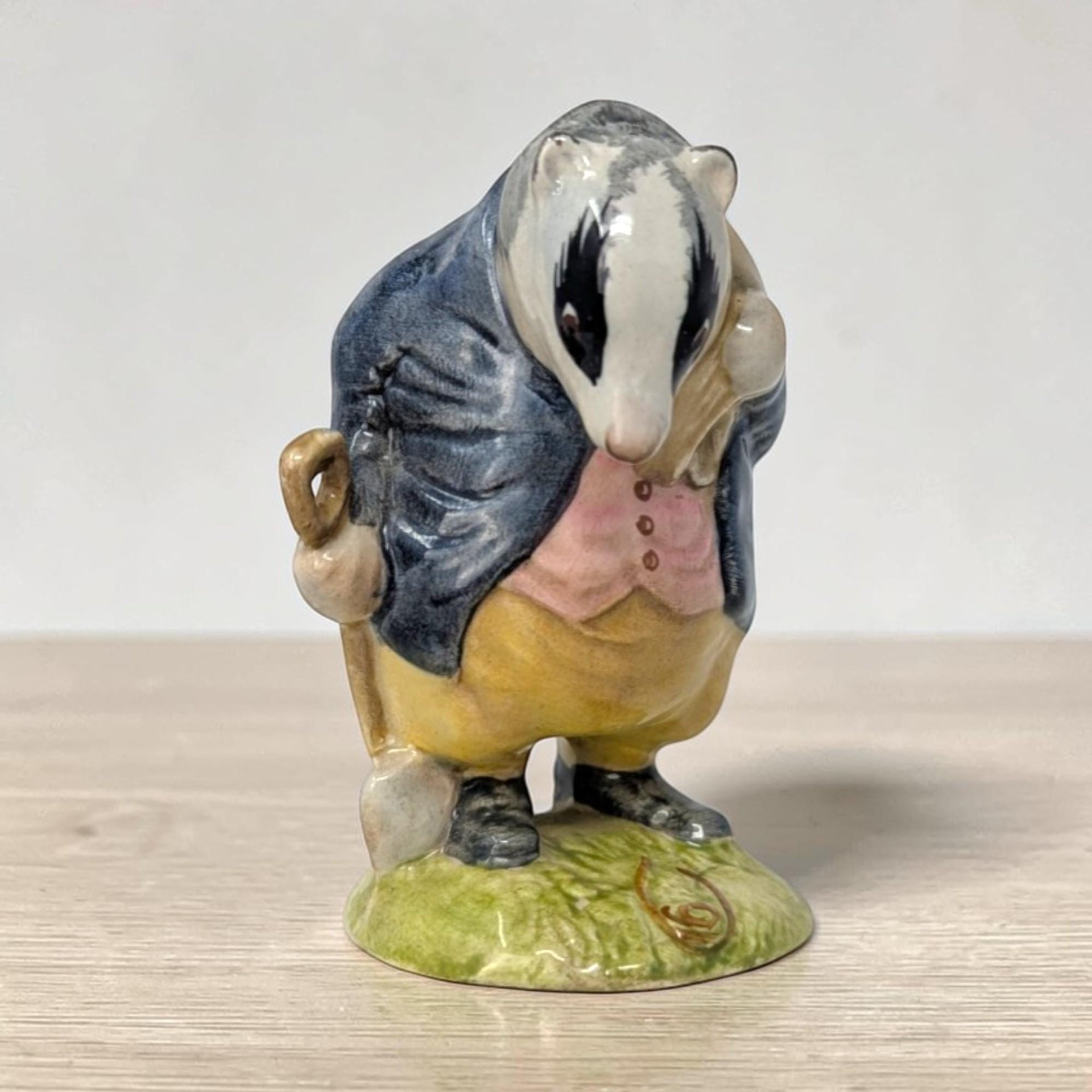 A small ceramic figurine of a badger character wearing a blue jacket over a  waistcoat. He holds a brown shovel and is finished in a high-gloss glaze with natural grey and white fur textures.A small ceramic figurine of a badger character wearing a blue jacket over a  waistcoat. He holds a brown shovel and is finished in a high-gloss glaze with natural grey and white fur textures.