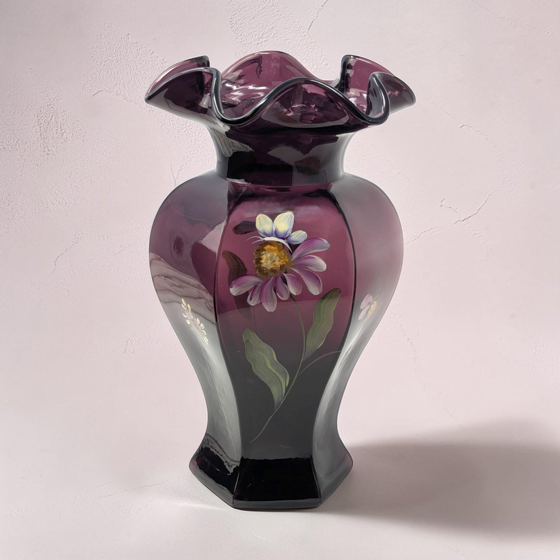 A tall, elegant vase in deep mulberry purple glass with a satin finish. It features hand-painted white floral sprigs and soft green leaves. The vase has a flared, ruffled rim and a rounded body.